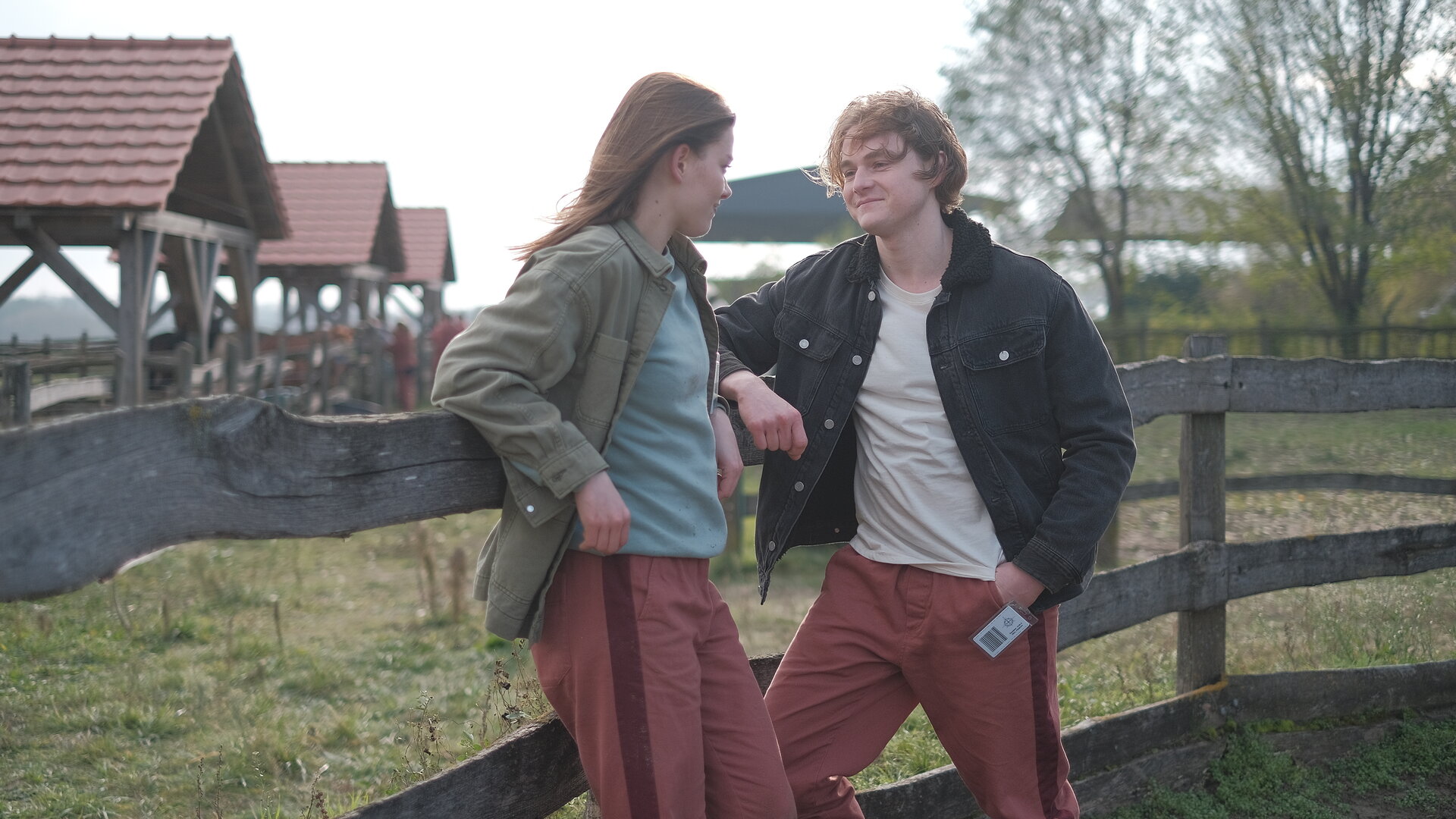 "Fallen - Unwetter": Luce (Jessica Alexander) und Cam (Timothy Innes) lehnen entspannt an einem hölzernen Zaun und blicken sich lächelnd an. Im Hintergrund stehen mehrere Stallgebäude und Bäume.