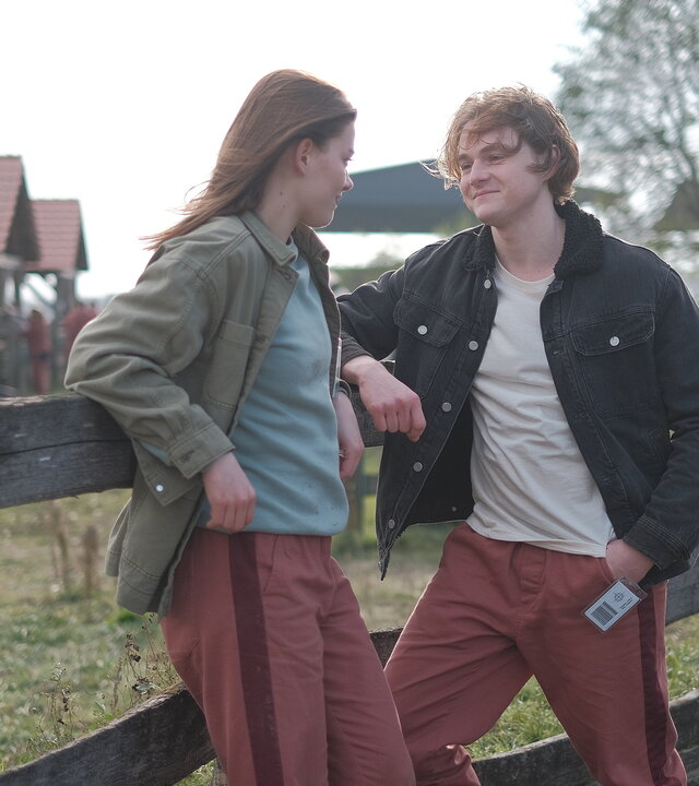 "Fallen - Unwetter": Luce (Jessica Alexander) und Cam (Timothy Innes) lehnen entspannt an einem hölzernen Zaun und blicken sich lächelnd an. Im Hintergrund stehen mehrere Stallgebäude und Bäume.