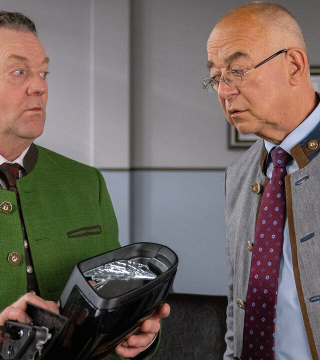 "Die Rosenheim-Cops - Spieglein, Spieglein": Bürgermeister Karl Schretzmayer (Jürgen Tonkel) und Polizeidirektor Achtziger (Alexander Duda) stehen in Achtzigers Büro und unterhalten sich. Schretzmayer hält einen schwarzen Außenspiegel in den Händen, den Achtziger mit prüfendem Blick betrachtet.