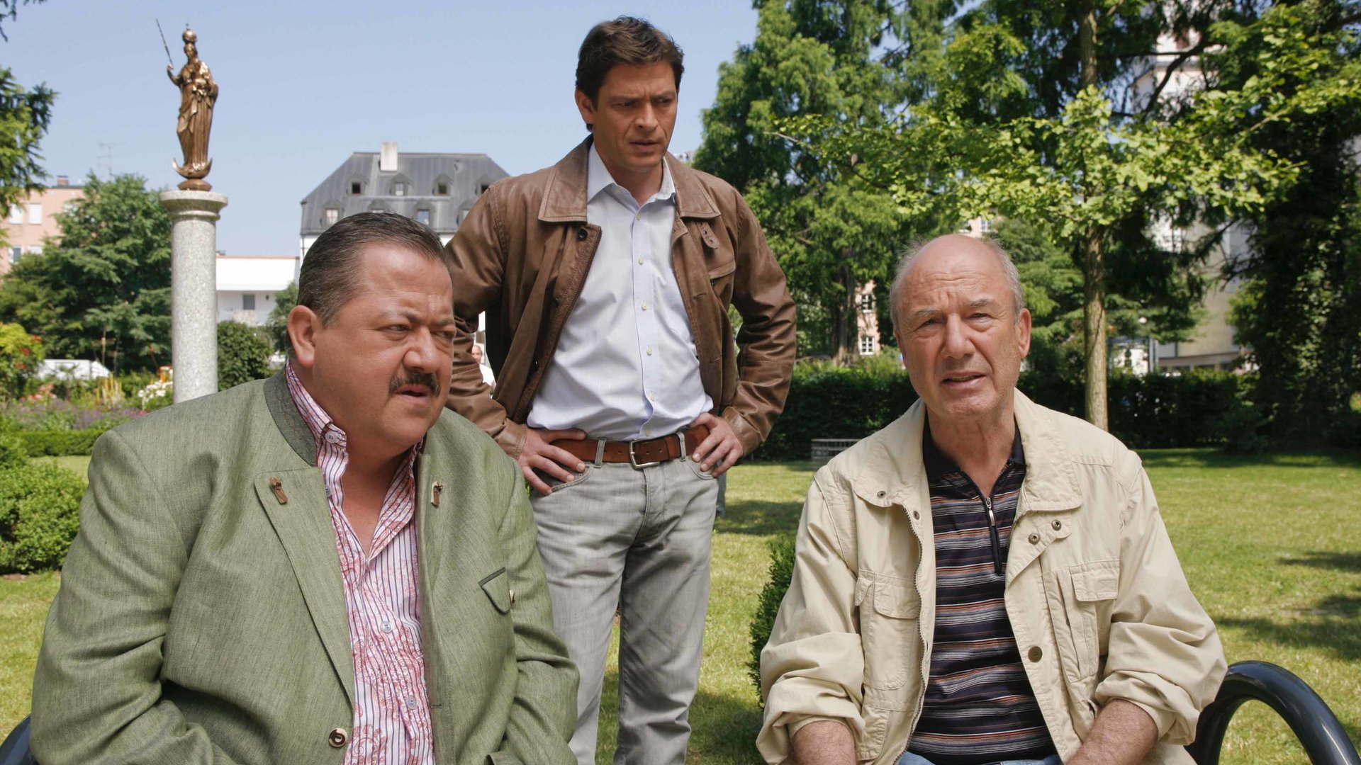 "Die Rosenheim-Cops - Tod beim Live-Chat": Korbinian Hofer (Joseph Hannesschläger) und Siegfried Dankl (Dietz-Werner Steck) sitzen nebeneinander auf einer Holzbank in einer Grünanlage und unterhalten sich. Hinter ihnen steht Christian Lind (Tom Mikulla), er hat die Hände in die Hüften gestützt. Im Hintergrund sind eine Bronzestatue auf einem hellen Steinsockel zu sehen sowie ein Haus und Bäume.