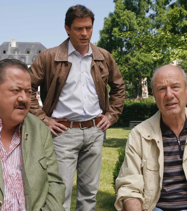 "Die Rosenheim-Cops - Tod beim Live-Chat": Korbinian Hofer (Joseph Hannesschläger) und Siegfried Dankl (Dietz-Werner Steck) sitzen nebeneinander auf einer Holzbank in einer Grünanlage und unterhalten sich. Hinter ihnen steht Christian Lind (Tom Mikulla), er hat die Hände in die Hüften gestützt. Im Hintergrund sind eine Bronzestatue auf einem hellen Steinsockel zu sehen sowie ein Haus und Bäume.