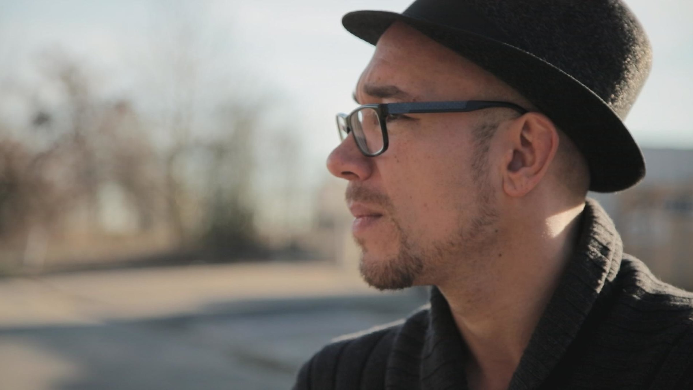 Besonders normal: "Aus der Bahn geworfen!": Auf der rechten Seite im seitlichen Profil einen Mann mit Hut und Brille, der in die Ferne schaut. Im Hintergrund verschwommene Bäume und einen Weg