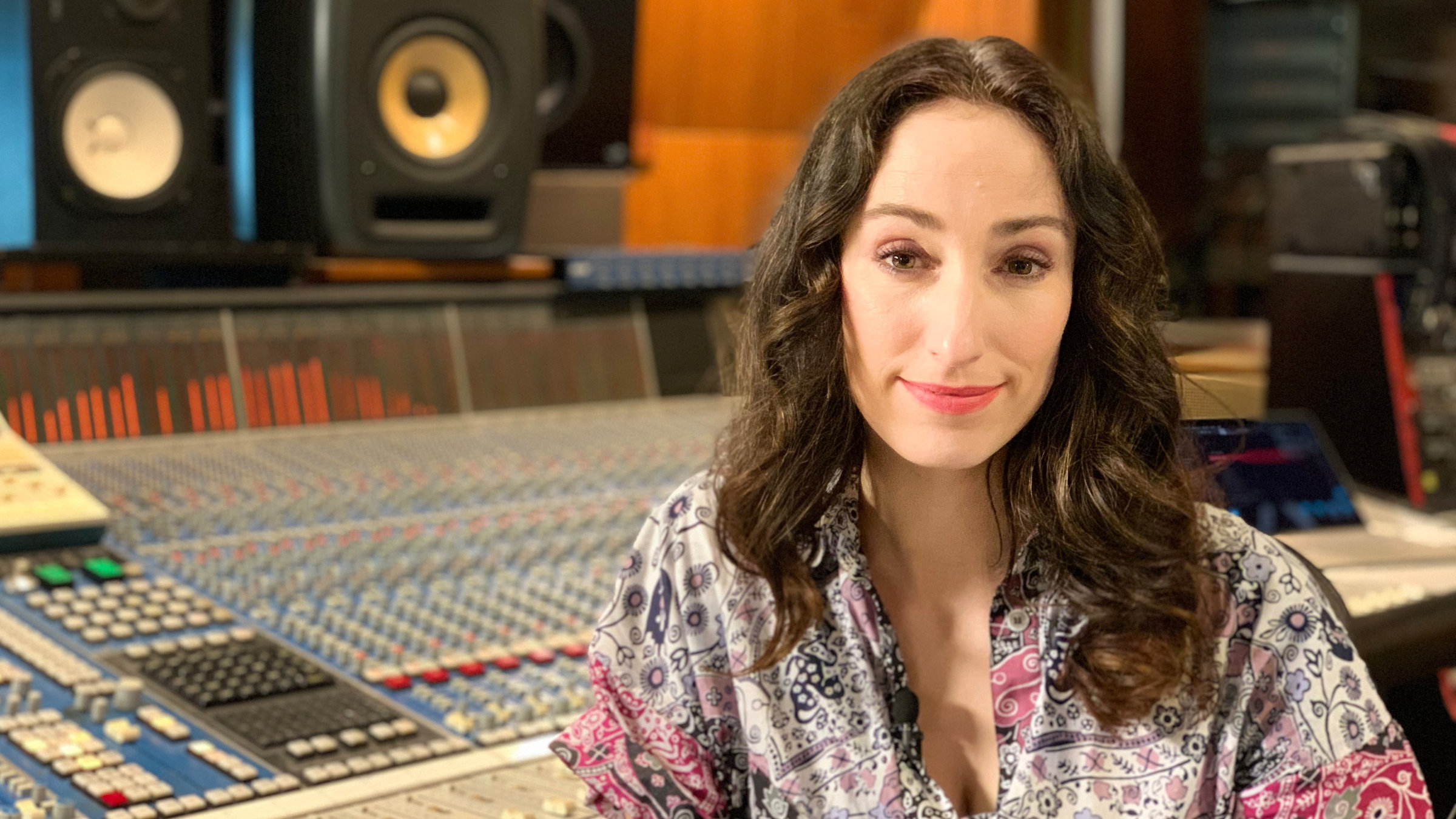 "Pop Around - The Video Stars": Eine Frau mit langen dunklen Locken sitzt in einem gemusterten Kleid vor einem großen Mischpult, im Hintergrund stehen zwei Boxen.