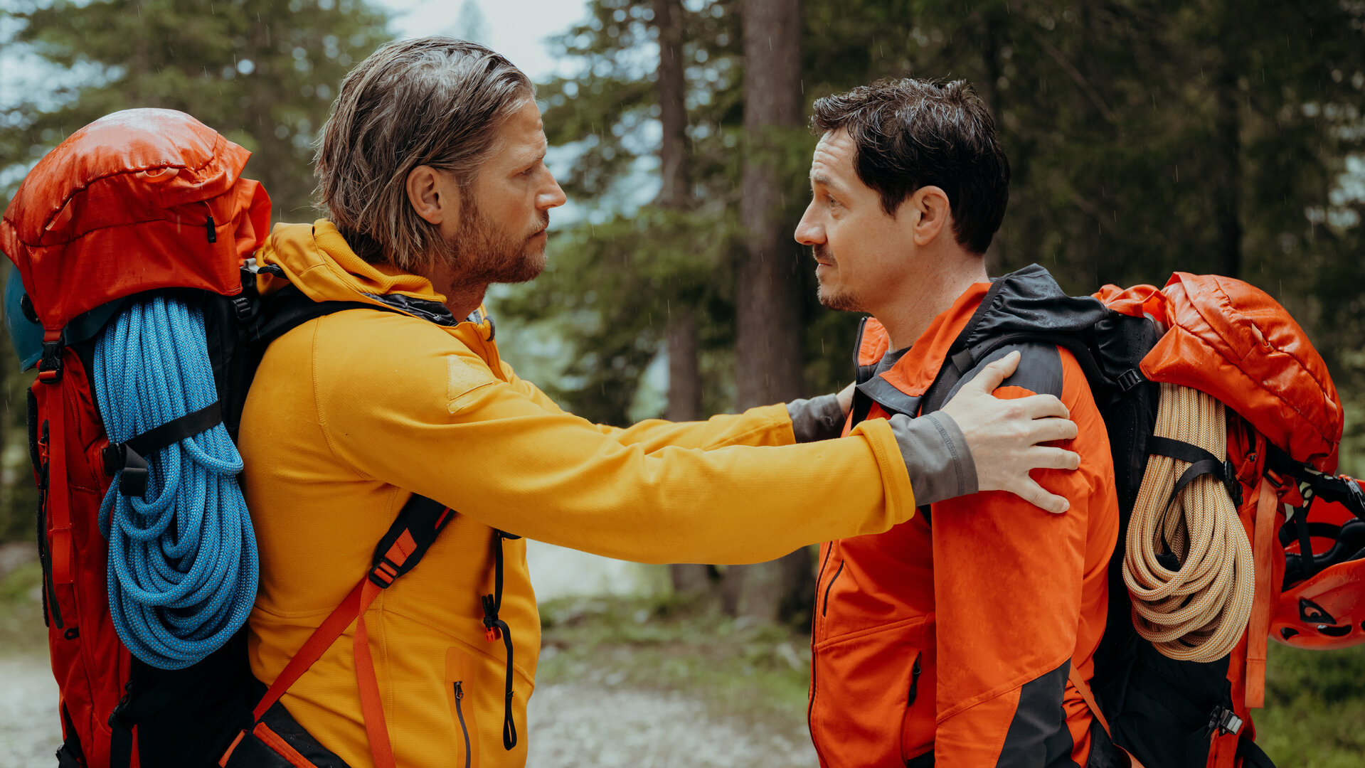 "Die Bergretter: Höhenfeuer (2)": Auf einem Waldweg: Die Bergretter Markus (Sebastian Ströbel) und Tobi (Markus Brandl) stehen sich gegenüber. Markus hat die Hände auf Tobis Arme gelegt und blickt ihn bittend an. Dieser hat die Stirn gerunzelt. Beide tragen Rucksäcke, an denen Seile und Helme befestigt sind.