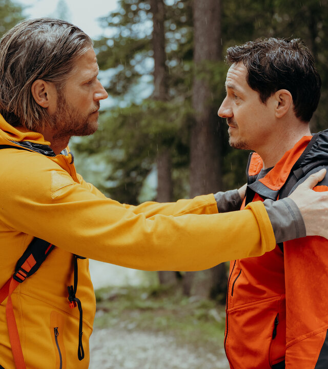 "Die Bergretter: Höhenfeuer (2)": Auf einem Waldweg: Die Bergretter Markus (Sebastian Ströbel) und Tobi (Markus Brandl) stehen sich gegenüber. Markus hat die Hände auf Tobis Arme gelegt und blickt ihn bittend an. Dieser hat die Stirn gerunzelt. Beide tragen Rucksäcke, an denen Seile und Helme befestigt sind.