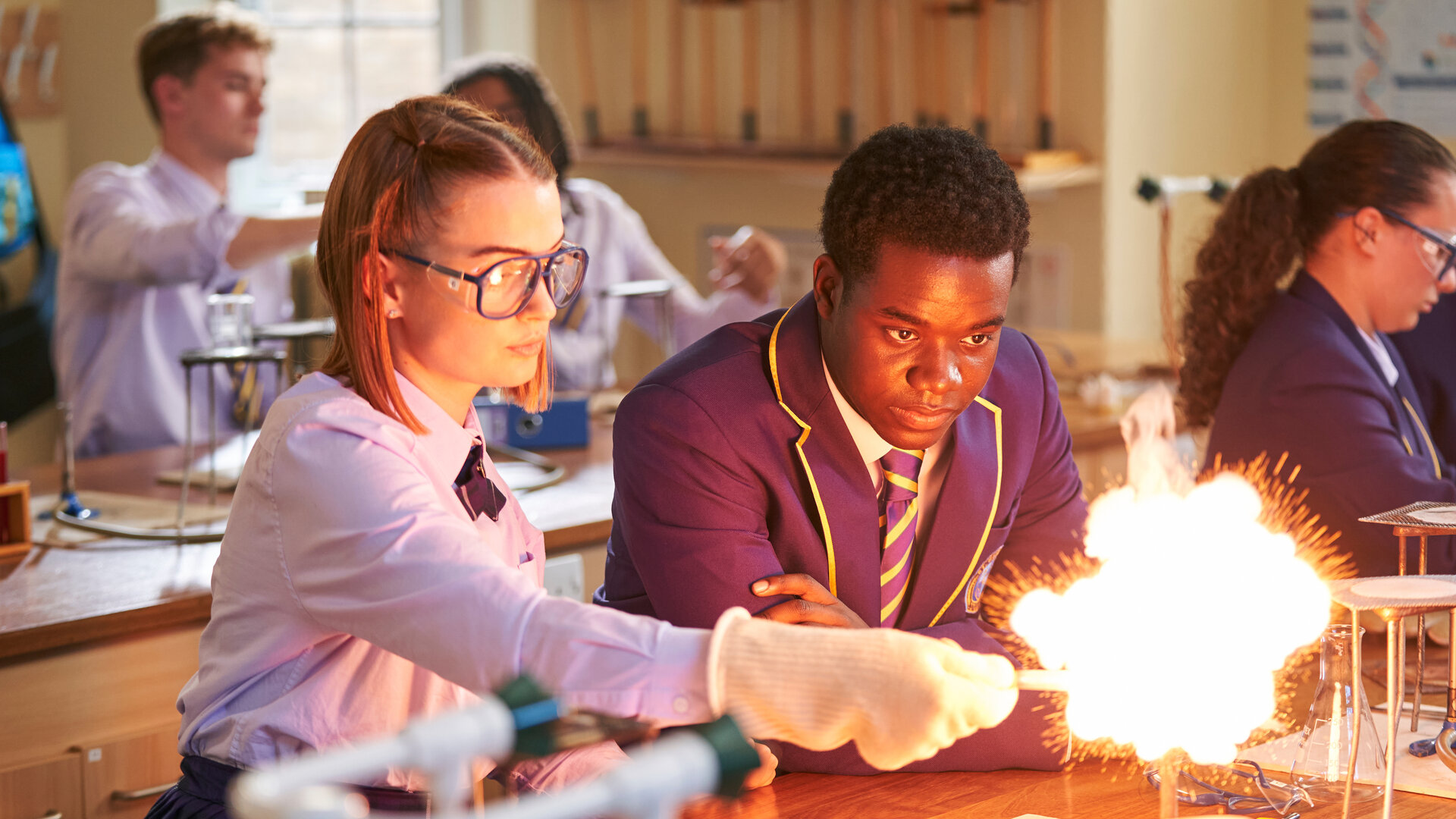 "Boarders - Welcome to St. Gilbert's - Dances & Decisions": Beatrix (Tallulah Greive) steht in einem Chemieraum und führt gerade ein Experiment durch, bei dem es zu einer Stichflamme kommt. Sie trägt eine Schutzbrille und Handschuhe. Jaheim (Josh Tedeku) steht neben ihr und blickt in die Flammen. Hinter ihnen sitzen weitere Schülerinnen und Schüler (Komparsen).
