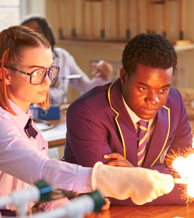 "Boarders - Welcome to St. Gilbert's - Dances & Decisions": Beatrix (Tallulah Greive) steht in einem Chemieraum und führt gerade ein Experiment durch, bei dem es zu einer Stichflamme kommt. Sie trägt eine Schutzbrille und Handschuhe. Jaheim (Josh Tedeku) steht neben ihr und blickt in die Flammen. Hinter ihnen sitzen weitere Schülerinnen und Schüler (Komparsen).