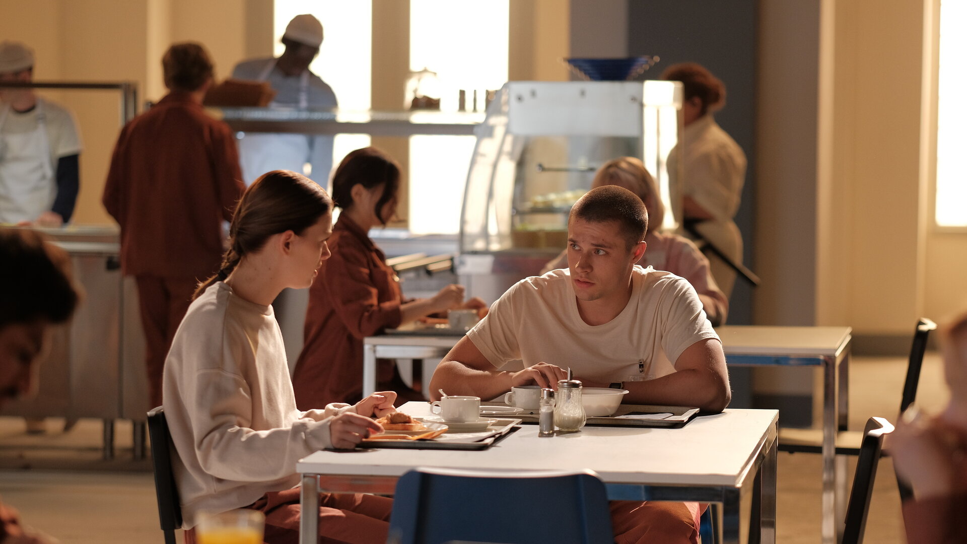 "Fallen - Schmetterlinge": Luce (Jessica Alexander) und Daniel (Gijs Blom) sitzen sich in einer Kantine an einem Tisch gegenüber und blicken sich ernst an. Vor ihnen stehen Tabletts mit Essen. Im Hintergrund sind weitere Personen (Komparsen) beim Essen und an der Essensausgabe zu sehen.