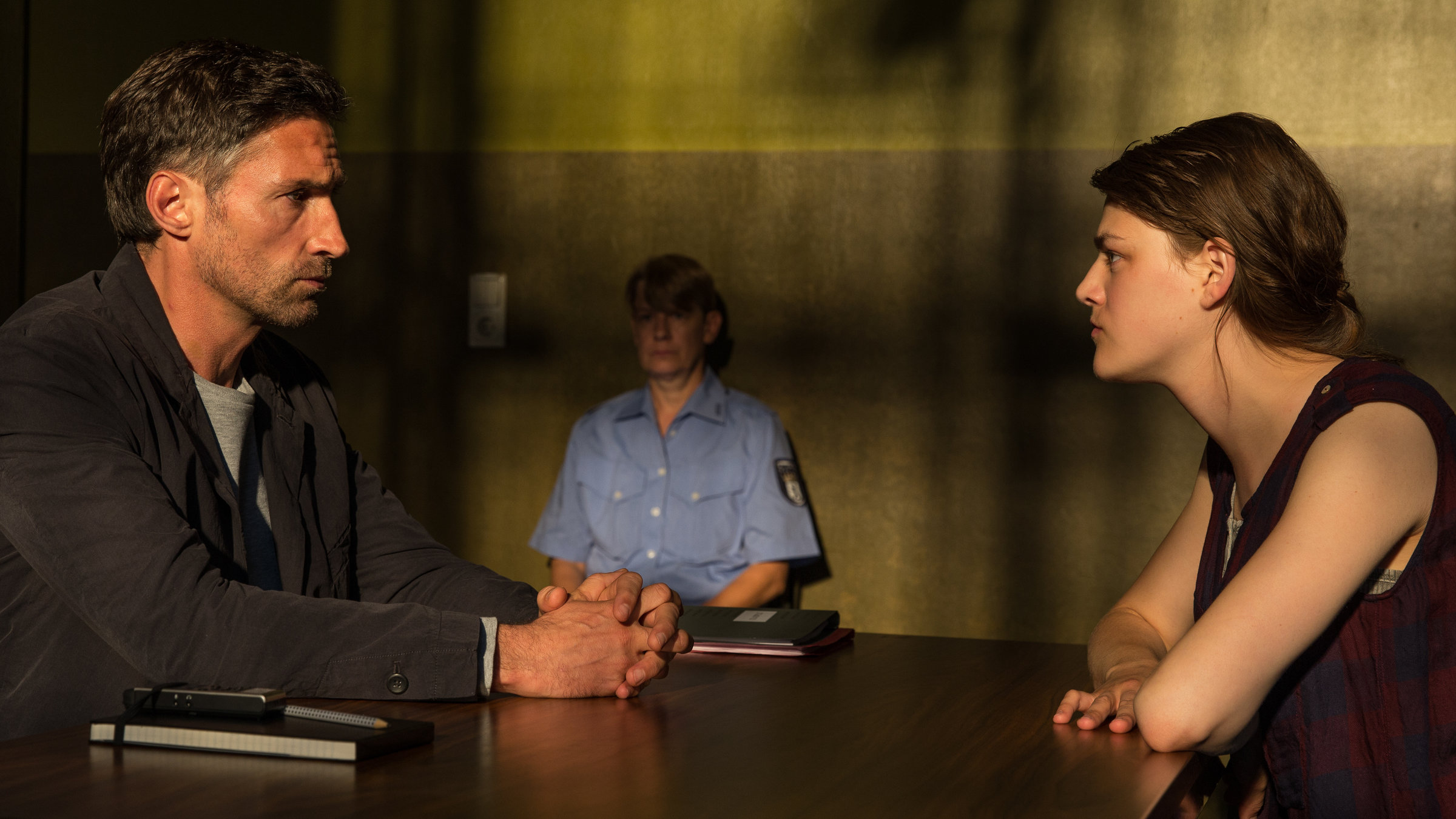 "Der Gutachter - Ein Mord zu viel": Robert Siedler (Benjamin Sadler) sitzt mit ernstem Gesichtsausdruck gegenüber Rebekka Lorenz (Johanna Polley) an einem Tisch. Die beiden schauen sich an. Im Hintergrund sitzt die Schließerin (Cornelia Lippert), die die Situation beobachtet.