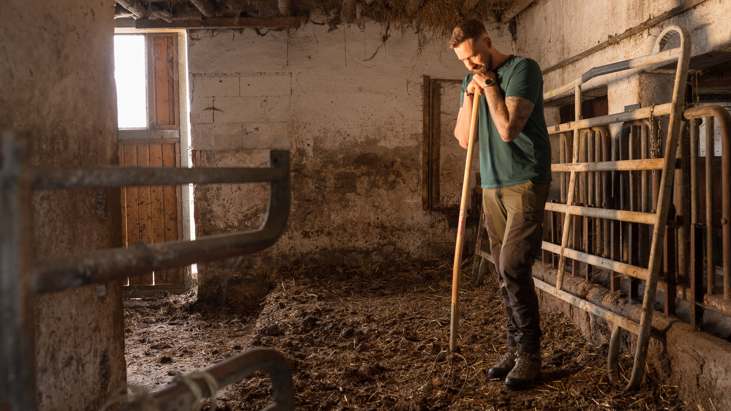 "37°: Burnout auf dem Bauernhof - Landwirte kämpfen gegen ihre Depression": Christoph Rothhaupt steht im Stall. Er ist bekleidet mit festem Schuhwerk, Outdoor-Hose und grünem Shirt. Sein linker Arm ist tätowiert. Er stützt sich auf einer Mistgabel ab und schaut mit nachdenklichem Blick zu Boden. Im Vordergrund sowie an der Wand stehen Gitter. Der Boden ist bedeckt mit Stroh und Gülle. Hinten ist eine Tür, von der Tageslicht von draußen hereinscheint.