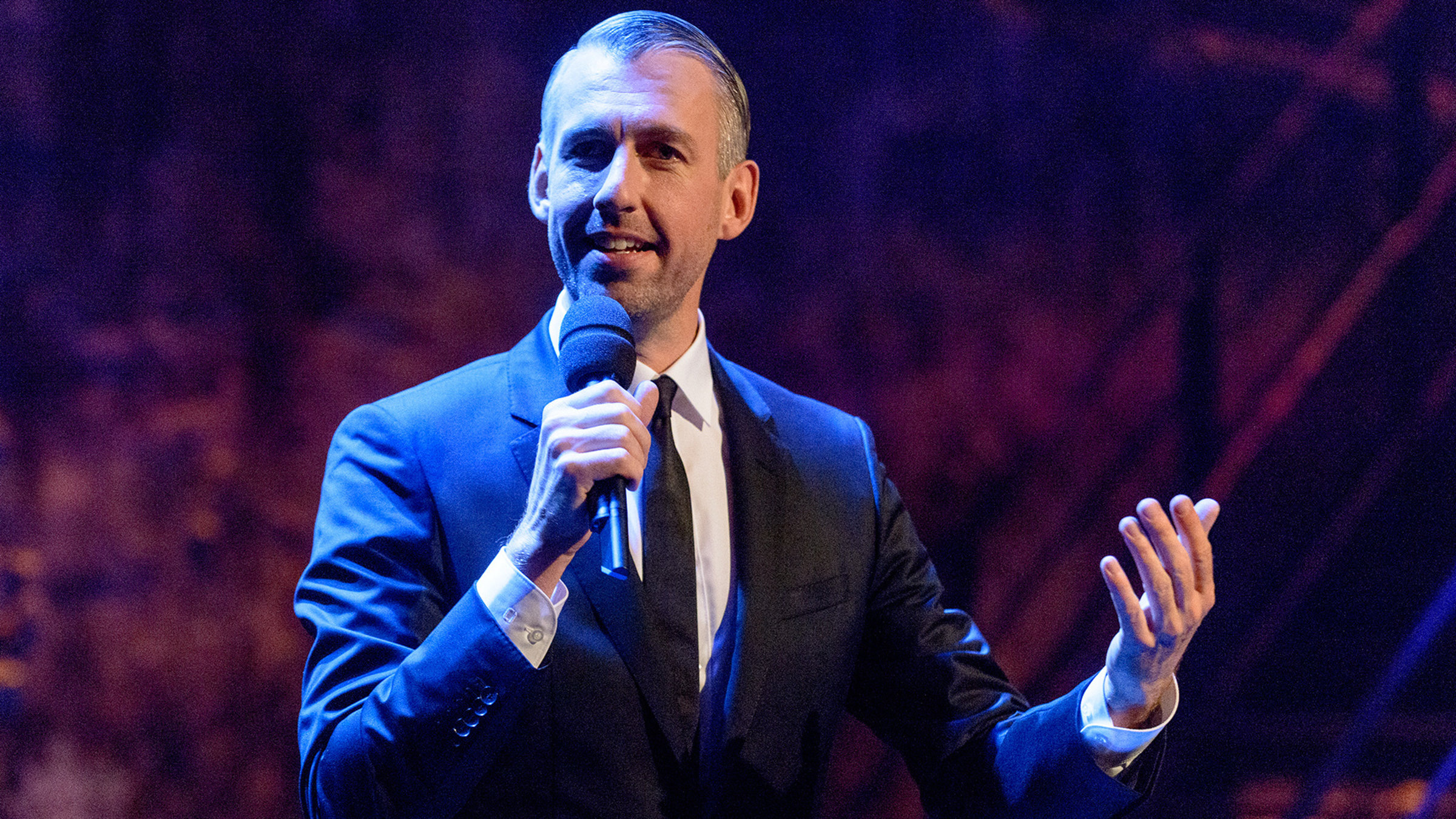 "Pufpaffs Happy Hour": Sebastian Pufpaff steht mit einem Mikrofon in der Hand auf der Bühne. Er trägt einem schwarzen Anzug mit weißem Hemd  und einer schwarzen Krawatte.