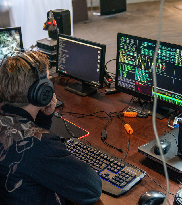 "The Bank Hacker": Andy (Joren Seldeslachts) und Jeremy (Tijmen Govaerts) sitzen gemeinsam vor mehreren Bildschirmen und blicken in diese hinein. Andy trägt Kopfhörer.