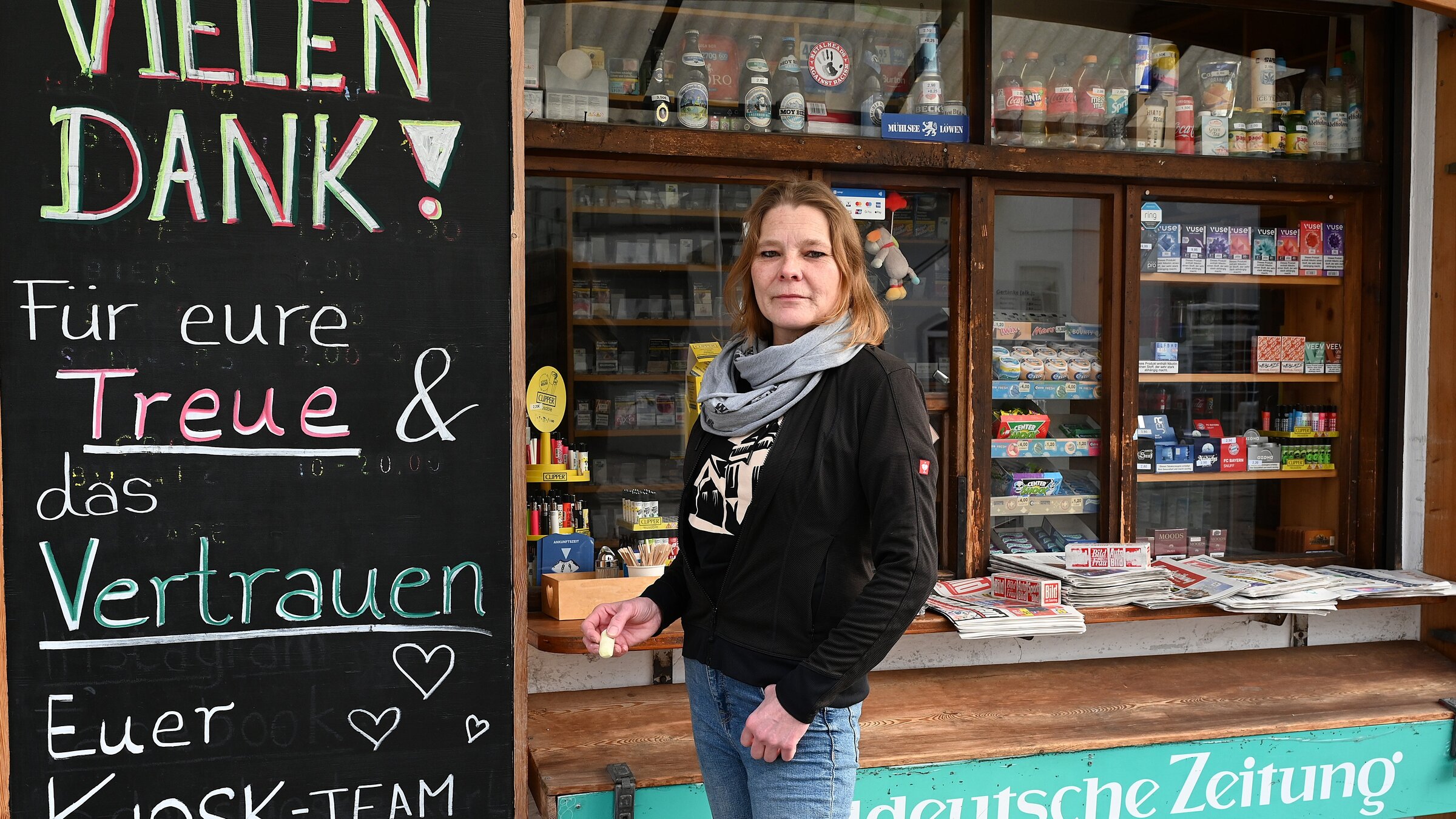 "ZDF.reportage: Selbstständige am Limit - Viel Arbeit für zu wenig Geld": Daniela E. steht vor ihrem Kiosk und schaut in die Kamera. Neben ihr ist eine Tafel zu sehen, auf der Abschiedsworte an ihre Kundschaft stehen.