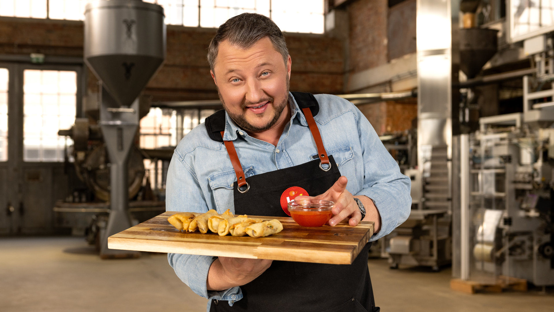 "ZDFbesseresser: Die Tricks der Asia-Restaurants - Sebastian Lege deckt auf": Produktentwickler Sebastian Lege trägt ein Holztablett. Darauf liegen sechs Frühlingsrollen. In seiner linken Hand hält er eine Glasschale mit süßsaurer Sauce.