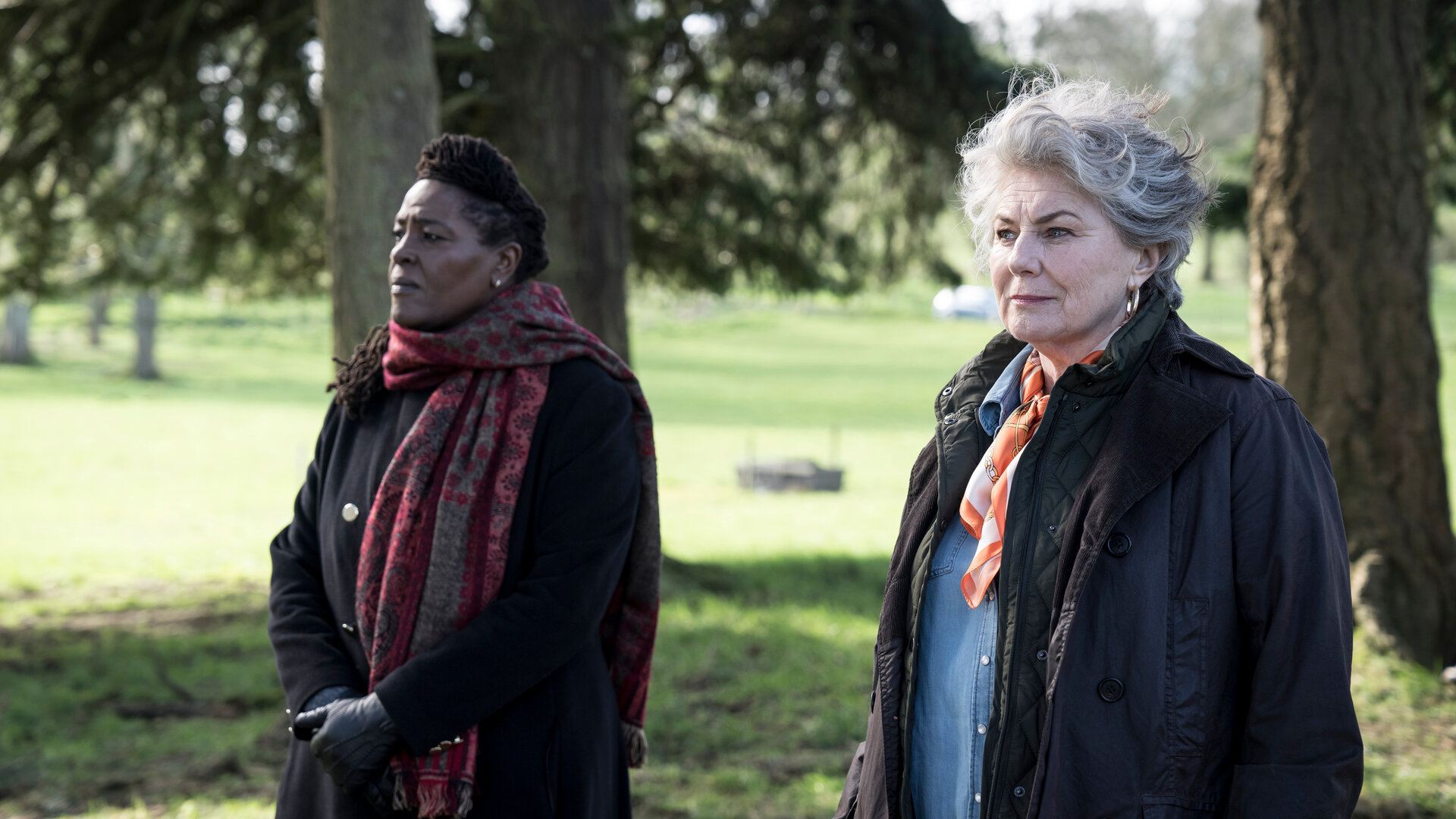 "Ellis - Familienbande": DCI Ellis (Sharon D Clarke und Carla Duffy (Orla Charlton) gehen nebeneinander unter Bäumen entlang. Sie tragen Kleidung, die vor Wind und Regen schützt und blicken seitlich in dieselbe Richtung.