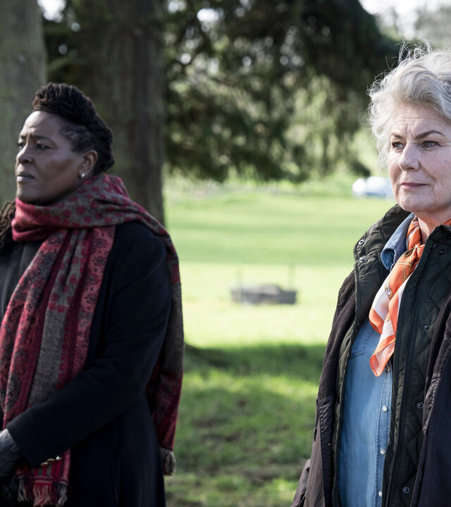 "Ellis - Familienbande": DCI Ellis (Sharon D Clarke und Carla Duffy (Orla Charlton) gehen nebeneinander unter Bäumen entlang. Sie tragen Kleidung, die vor Wind und Regen schützt und blicken seitlich in dieselbe Richtung.