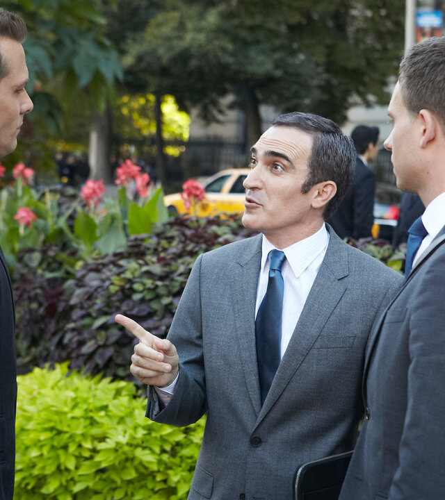 "Suits - Spannungsfelder": Harvey (Gabriel Macht) und Mike (Patrick J. Adams) stehen mit Elliott Stemple (Patrick Fischler) auf dem Gehweg vor einem Gebäude. Elliott Stemple (Patrick Fischler) zeigt gestikulierend auf Harvey. Hinter ihnen ist ein Beet mit Grünpflanzen, am Straßenrand stehen Bäume und Passanten (Komparsen) laufen die Straße mit parkenden Autos entlang.