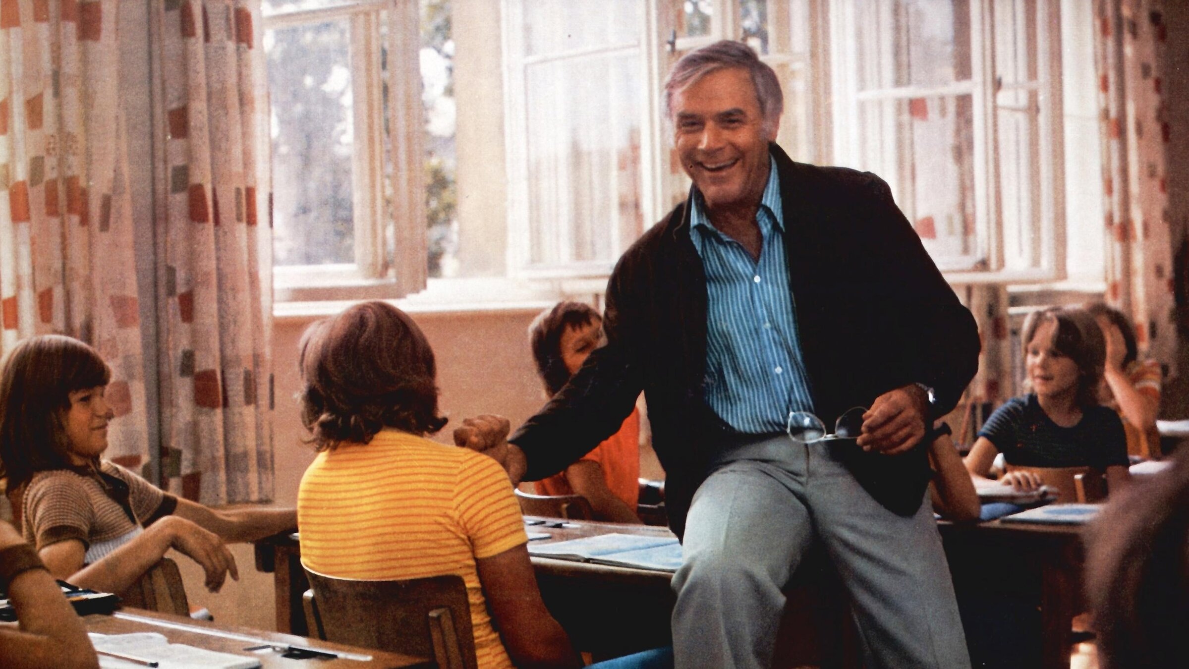 "Das fliegende Klassenzimmer": Joachim Fuchsberger als Lehrer Justus sitzt im Klassenzimmer locker und lachend auf dem Tisch eines Schülers, den er spaßeshalber am Handgelenk festhält. Justus trägt eine graue Hose, ein blau gestreiftes Hemd und einen dunklen Blouson.