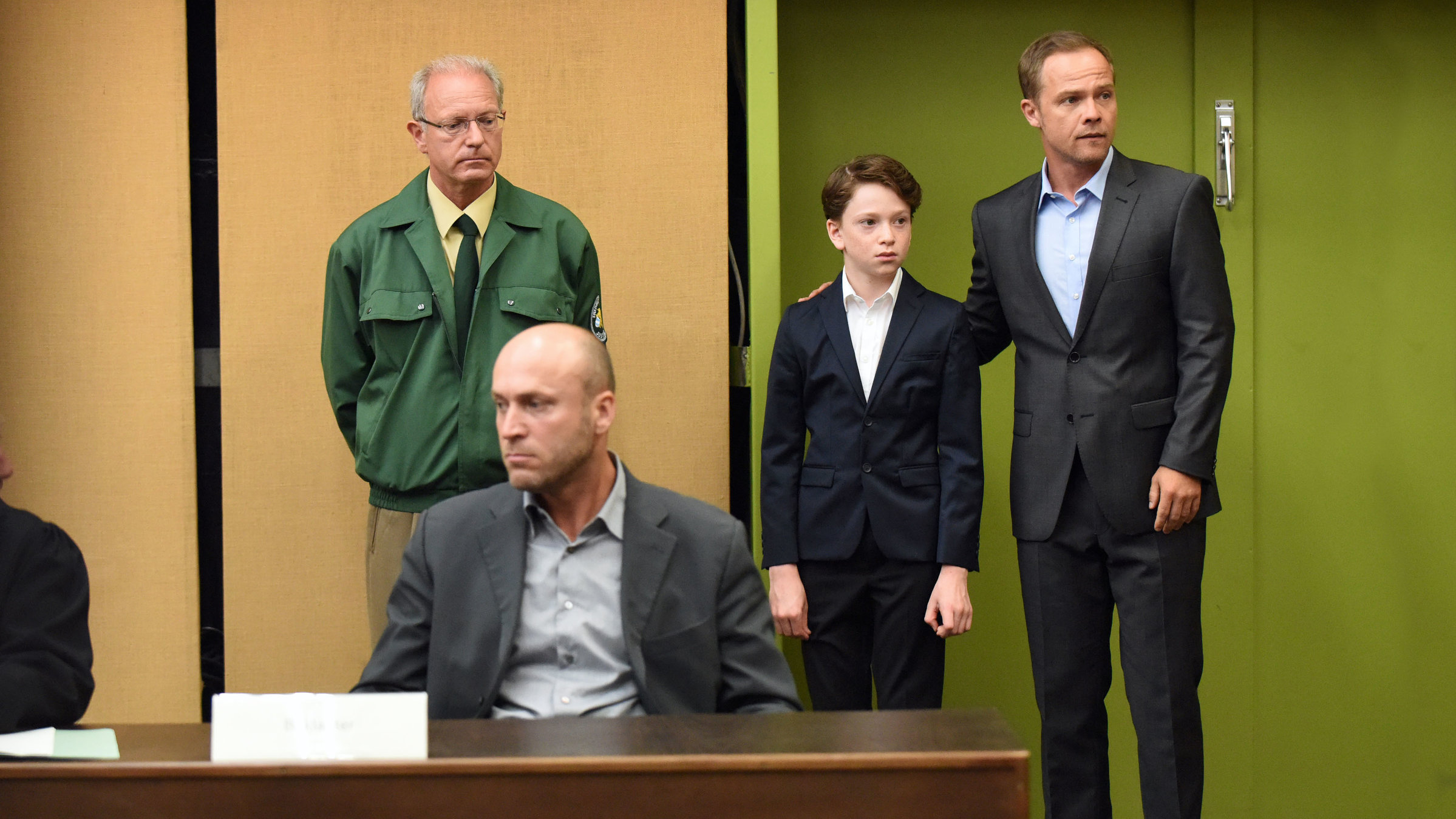 "Der Polizist, der Mord und das Kind": In einem Gerichtssaal sitzt Alexanders Vater (Ilir Rexhepi) am Tisch. Hinter ihm stehen Alexander (Joshio Marlon) und Opferschutzkommissar Carlos Benede (Matthias Koeberlin). Benede hat seinen rechten Arm um Alexanders Schulter gelegt.