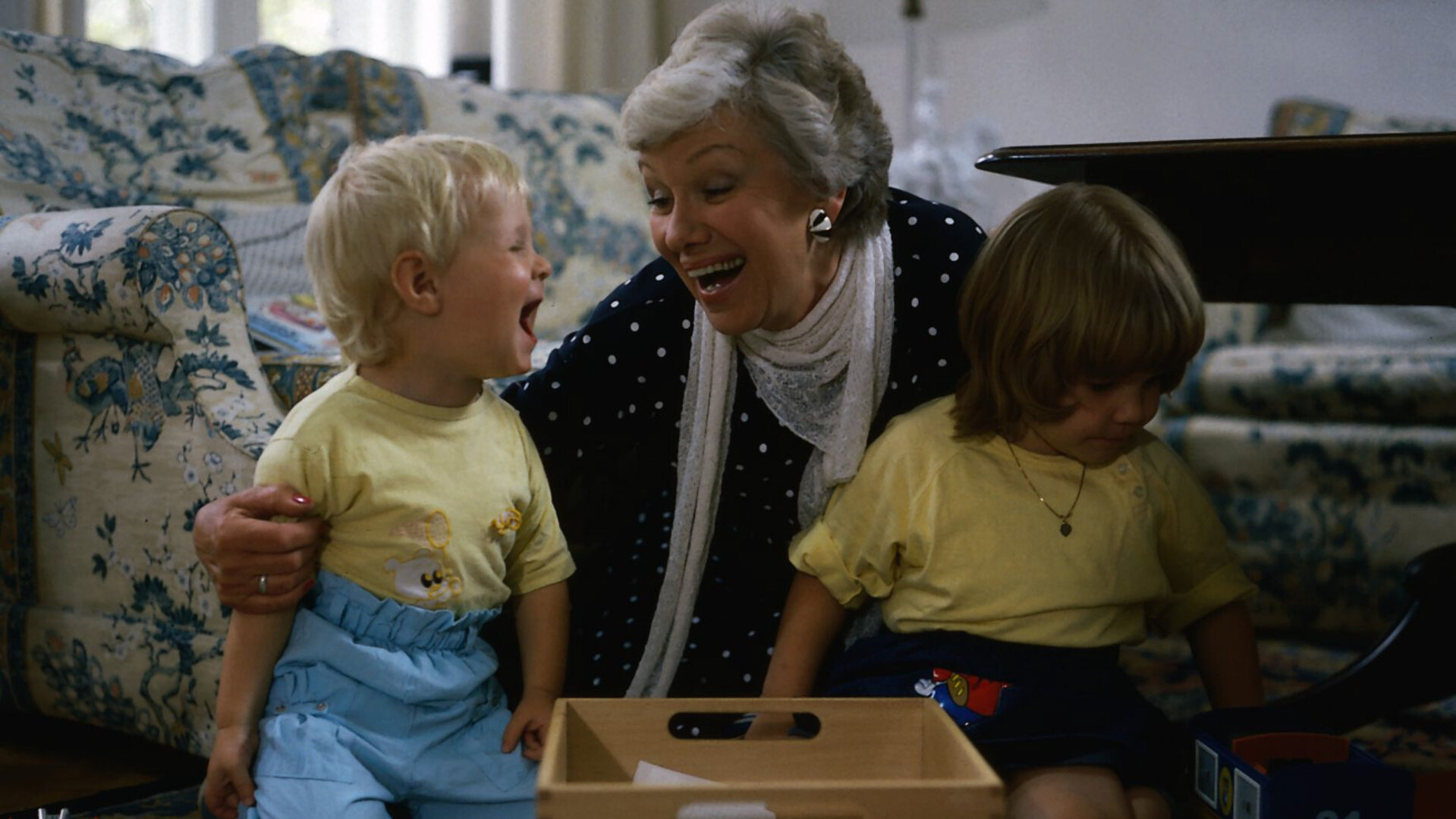 "Die Wicherts von nebenan: Die Heide, Hannelore und Hotels": Hannelore (Maria Sebaldt) sitzt im Wohnzimmer vor der Couch zwischen ihren Enkelkindern Sebastian (Oliver Lausberg) und Klein-Katrinchen (Sophie Birkner) und hat ganz viel Spaß.