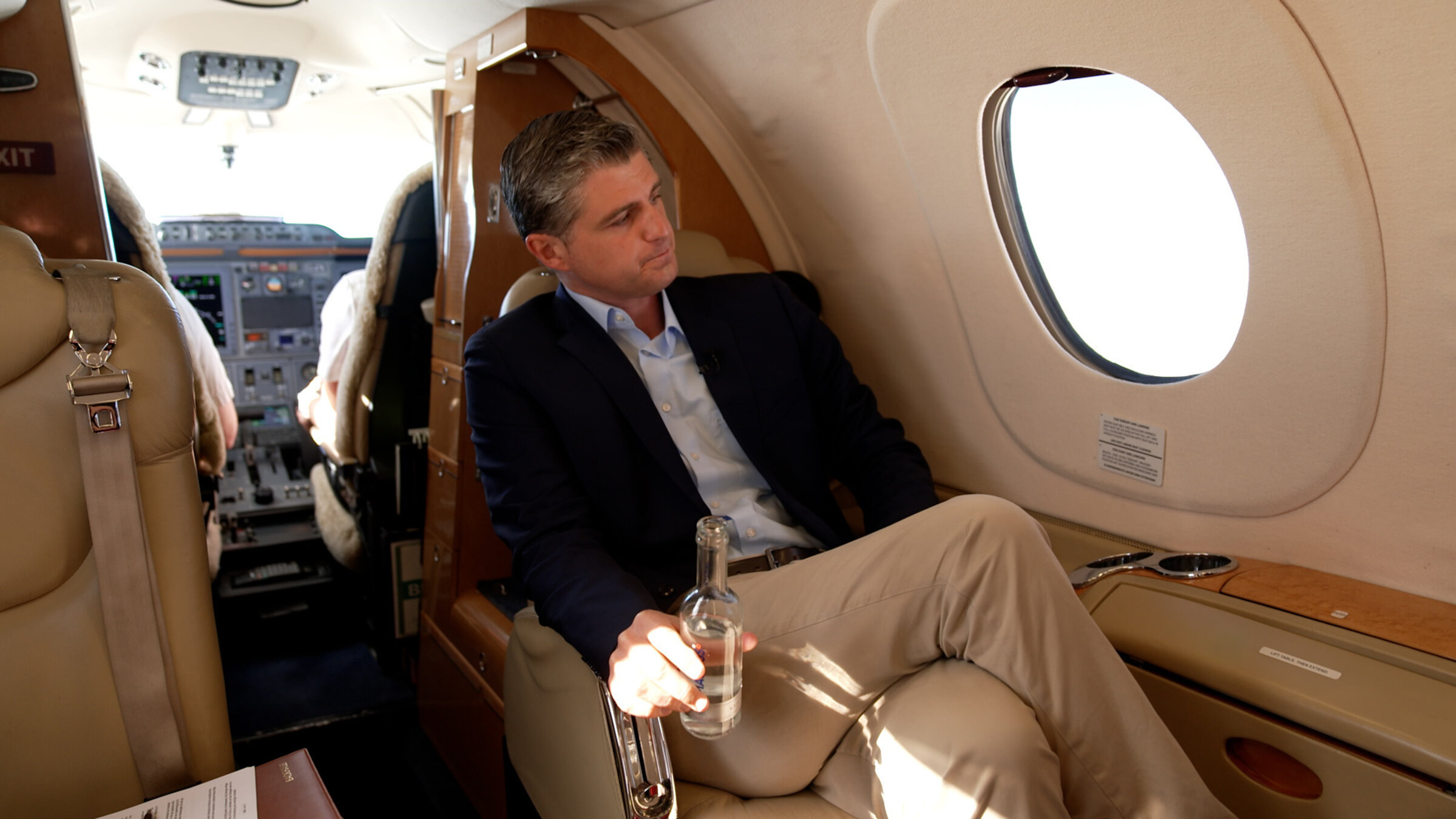 "ZDF.reportage: Luxus-VerkÃ¤ufer in Deutschland - Wie Reiche shoppen": Julien Backhaus sitzt mit Ã¼berschlagenen Beinen in einem Privatjet mit einer Wasserflasche in der Hand. Er schaut aus dem Fenster. Im Hintergrund sind die Schultern von Pilot und Co-Pilot im Cockpit zu sehen.