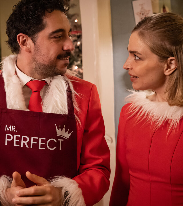 "Single Bells - Only Losers for Annie? - Damien (II)": Roel (Robbert Vervloet) und Mina (Julie Van den Steen) stehen in ihren vom Weihnachtsmann inspirierten Anzug und Kleid nebeneinander. Sie schauen sich intensiv an.