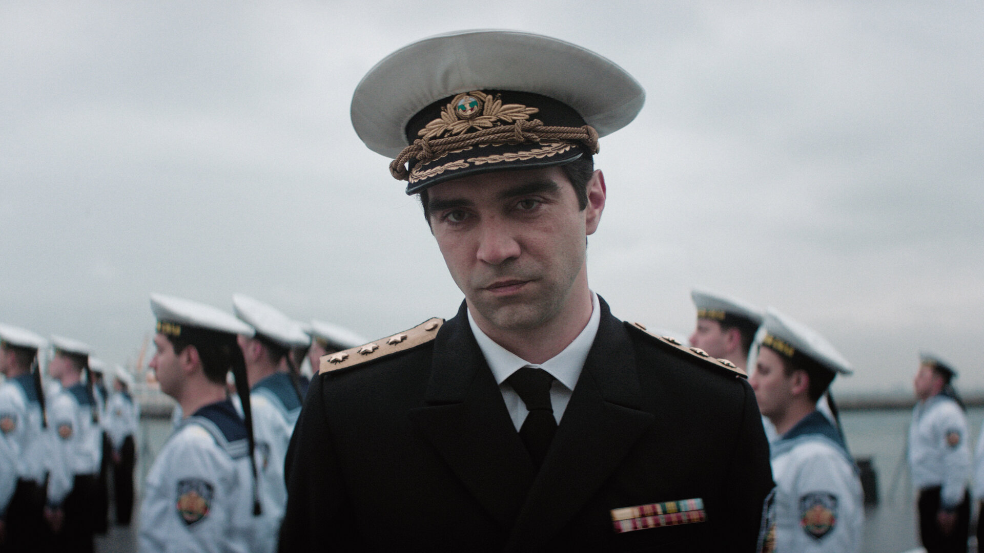 "The Veil - Das Asset": Emir (Alec Secăreanu) steht im Freien. Er trägt eine Offiziersuniform der Marine mitsamt Schirmmütze. Im Hintergrund steht eine Gruppe von Matrosen in Reihen.