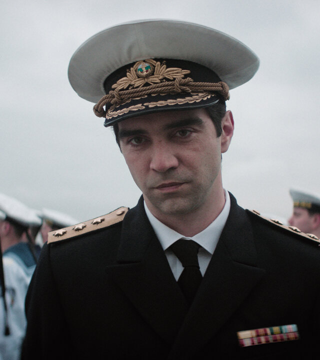 "The Veil - Das Asset": Emir (Alec Secăreanu) steht im Freien. Er trägt eine Offiziersuniform der Marine mitsamt Schirmmütze. Im Hintergrund steht eine Gruppe von Matrosen in Reihen.