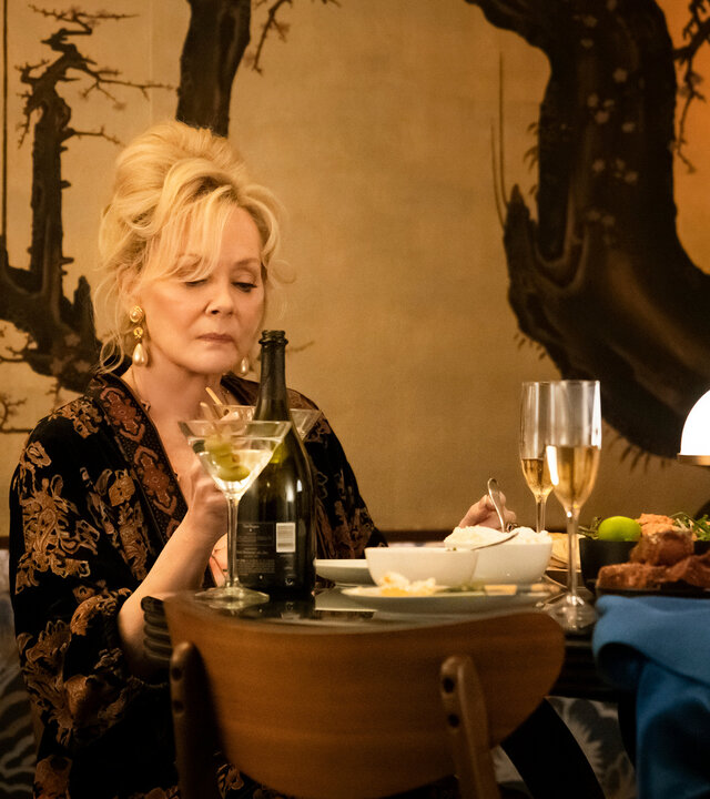 "Hacks - Ausschweifungen, hier und da": Deborah (Jean Smart) sitzt alleine an einem elegant gedeckten Tisch und hält ein Martini-Glas in der Hand, auf das sie nachdenklich blickt. Hinter ihr befindet sich eine kunstvoll gestaltete Wand mit einem baumartigen Muster.