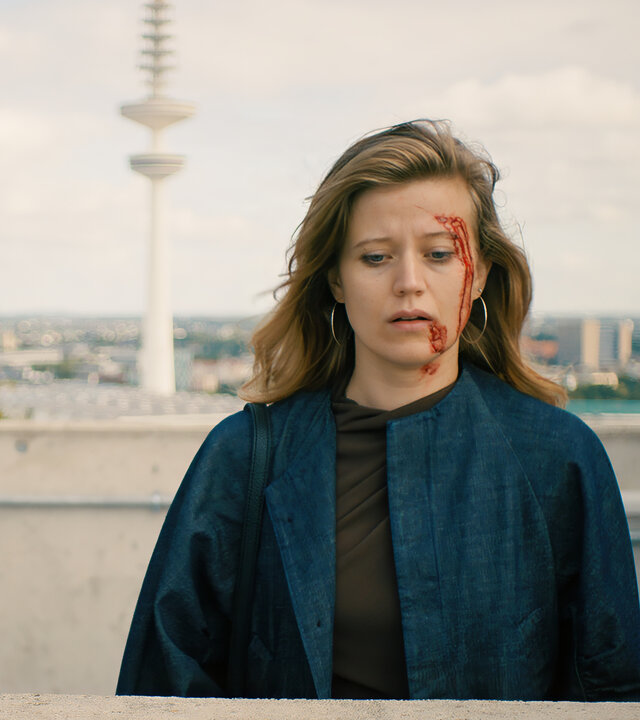 "Notruf Hafenkante - Dunkelrot": Milena Harnisch (Camille Dombrowsky) steht auf einer Dachterrasse. Sie blickt nachdenklich. Auf ihrer linken GesichtshÃ¤lfte befinden sich Blutspuren. Im Hintergrund liegt das Stadtpanorama von Berlin mit dem Funkturm.