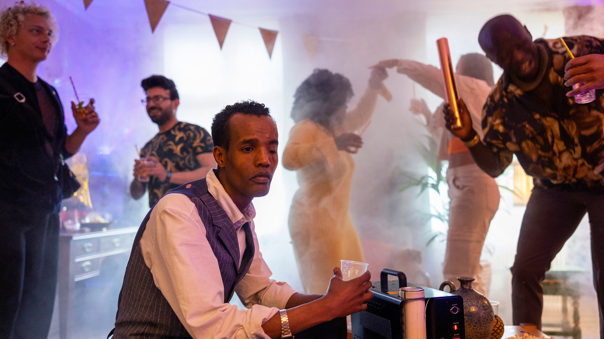 "Detective No. 24 - I Want to Know What Love Is": Ibraahin (Nasir Farah Dhagole) sitzt an einem Tisch und hält ein Getränk in der Hand. Im Hintergrund tanzen Latif (Malick Afocozi) und weitere Menschen. Der Raum ist mit Party-Utensilien geschmückt.