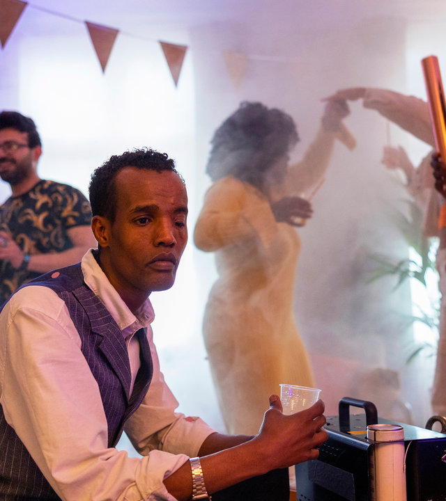 "Detective No. 24 - I Want to Know What Love Is": Ibraahin (Nasir Farah Dhagole) sitzt an einem Tisch und hält ein Getränk in der Hand. Im Hintergrund tanzen Latif (Malick Afocozi) und weitere Menschen. Der Raum ist mit Party-Utensilien geschmückt.