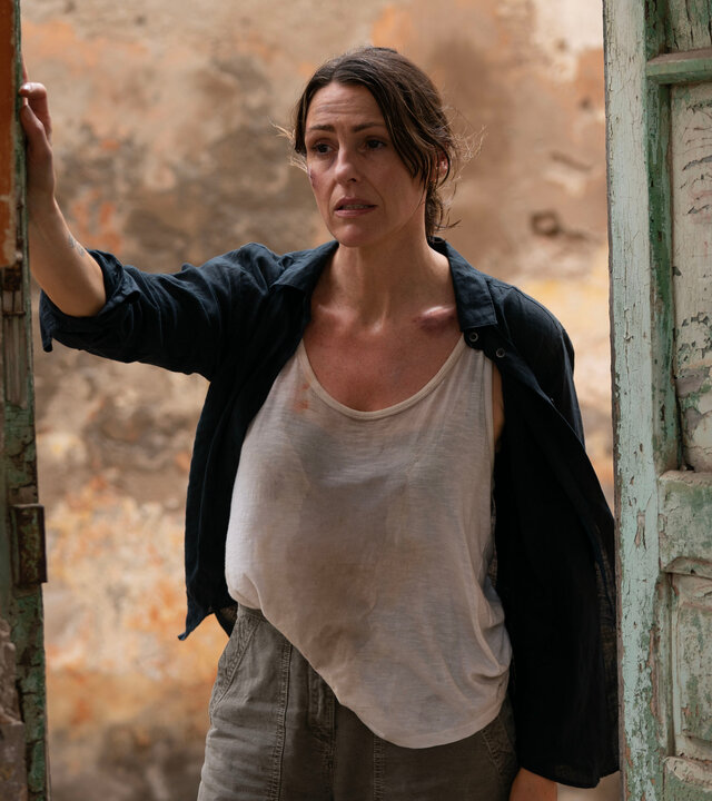 "Vigil - Tödliche Drohnen: Todesfalle": DCI Amy Silva (Suranne Jones) steht in einem alten Türrahmen, dessen grünlich-weiße Farbe abblättert. Sie trägt einfache Alltagskleidung, ihr weißes T-Shirt ist durchgeschwitzt. Sie blickt fragend während ihre rechte Hand auf der Wand links vom Türrahmen liegt.