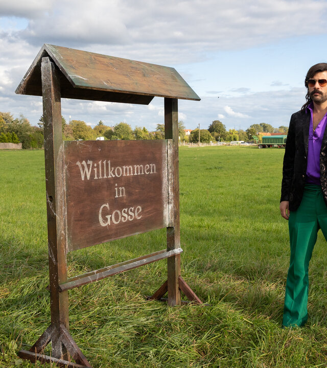 "Rembetis - die Geisterjäger - Who You Gonna Call": Paris (Jasin Challah) steht auf einer offenen Wiese vor einem Willkommensschild aus Holz seines Heimatdorfes Gosse.