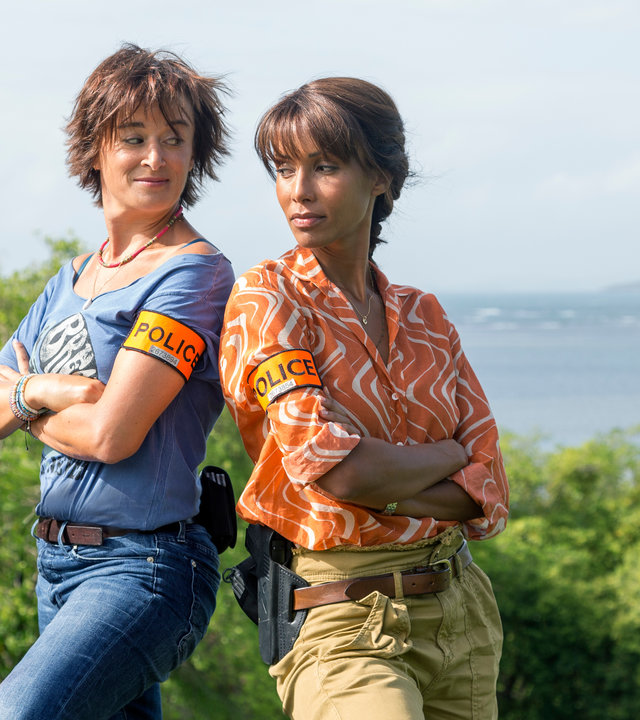 "Deadly Tropics - Opfer oder Täter?": Gaëlle Crivelli (Béatrice de la Boulaye) und Mélissa Sainte-Rose (Sonia Rolland) stehen Rücken an Rücken vor einer bewaldeten Klippe und schauen sich fröhlich an. Im Hintergrund ist das Meer zu erkennen.