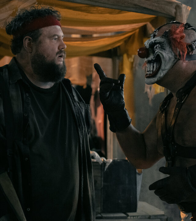 "Twisted Metal - LZGTBZY": Vor einem Verkaufsstand in einer Halle stehen sich Stu (Mike Mitchell) und Sweet Tooth (Joe Seanoa) gegenüber. Stu blickt mit leicht geöffnetem Mund zu Sweet Tooth fragend an. Sweet Tooth trägt eine Clownsmaske mit einem breiten Lächeln und zeigt mit einer Hand nach oben. Im Hintergrund befinden sich zwei Verkaufsstände, an denen Waffen und Munition angeboten werden.