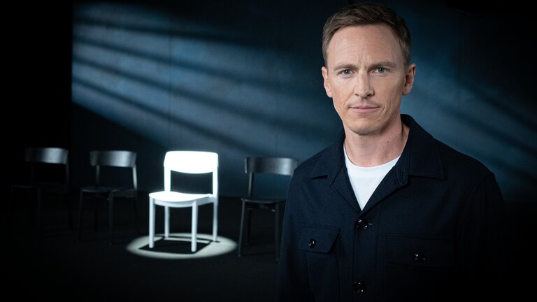 "Weil du mir gehörst! - Wenn Männer ihre Frauen töten": Jochen Breyer steht in einem abgedunkelten Raum und schaut in die Kamera. Im Hintergrund stehen mehrere Stühle, ein weißer Stuhl wird angestrahlt.