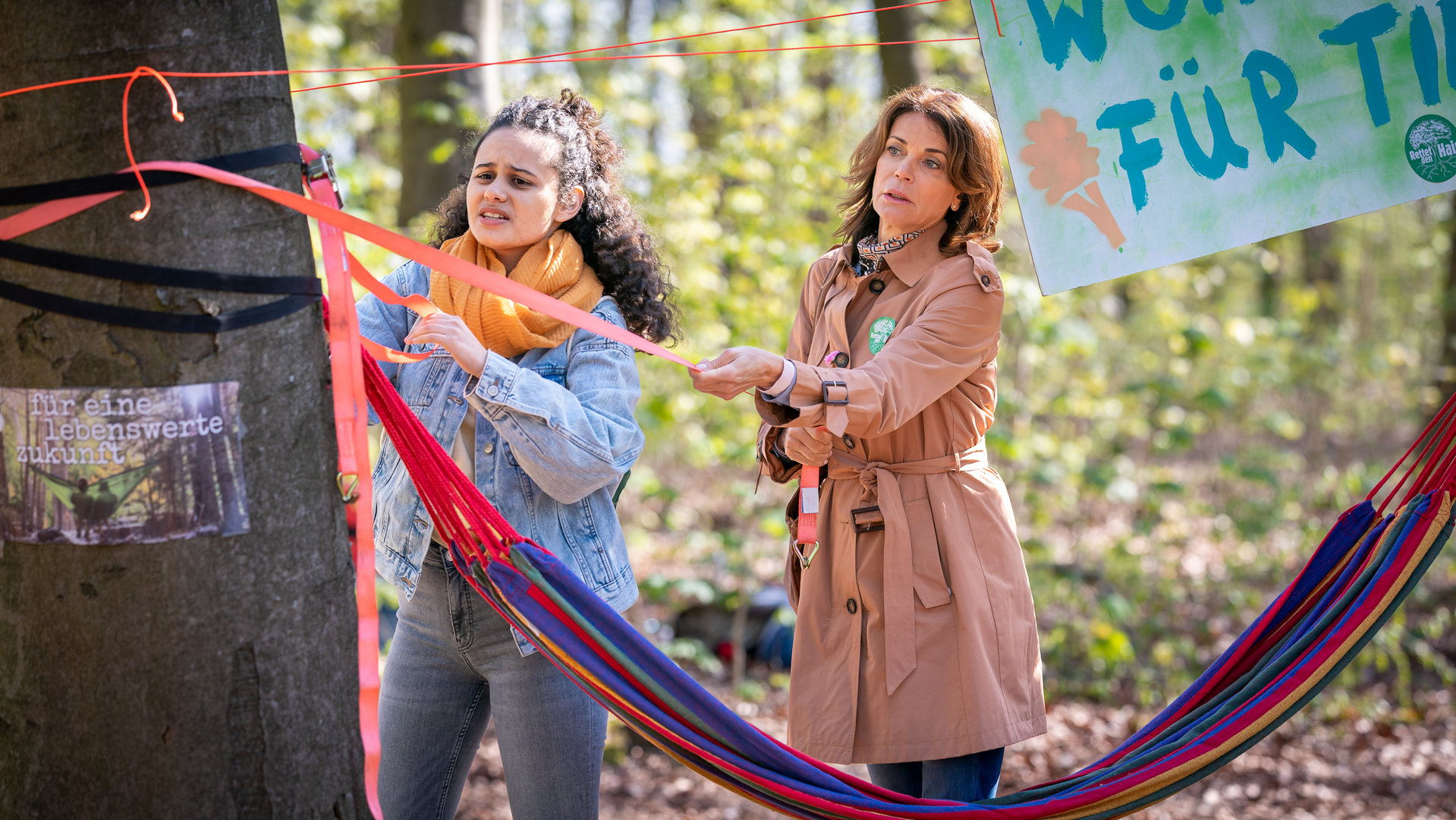 "Notruf Hafenkante - Eingesperrt": Kira (Saron Degineh) und Jasmin (Gerit Kling) befinden sich in einem Wald. Sie stehen neben einem Baumstamm und sind im Begriff ein Transparent mit Gurten zu befestigen.