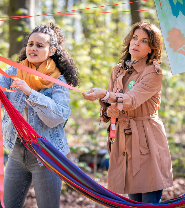 "Notruf Hafenkante - Eingesperrt": Kira (Saron Degineh) und Jasmin (Gerit Kling) befinden sich in einem Wald. Sie stehen neben einem Baumstamm und sind im Begriff ein Transparent mit Gurten zu befestigen.