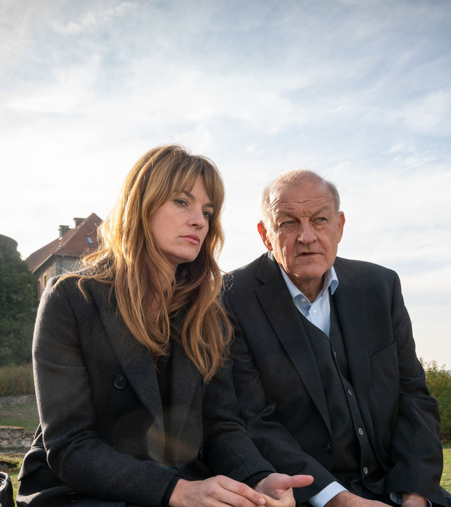 "Wilsberg - Ins Gesicht geschrieben":  Alex Holtkamp (Ina Paule Klink) sitzt mit traurigem Blick neben Georg Wilsberg (Leonard Lansink) auf einer Mauer. Im Hintergrund sieht man einen Turm der Sparrenburg.