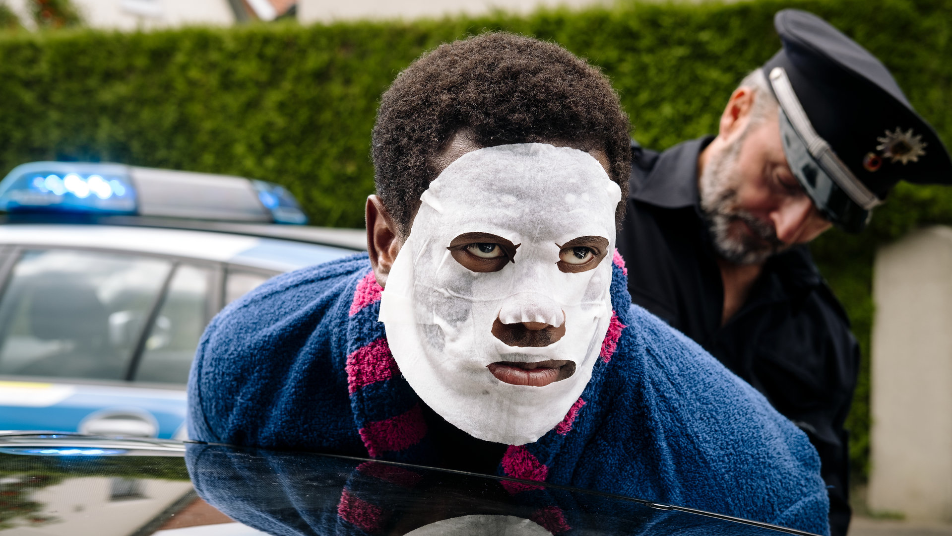 "I don't work here - Hecke": Dawit (Akeem van Flodrop) wird von Polizist Jakob (Robert Schupp) festgenommen. Während Jakob ihn in Polizeiuniform an der Autotür fixiert, lehnt Dawit seinen Oberkörper an ein dunkles Autodach und trägt eine weiße Tuchmaske und einen blauen Bademantel mit pinken Streifen.