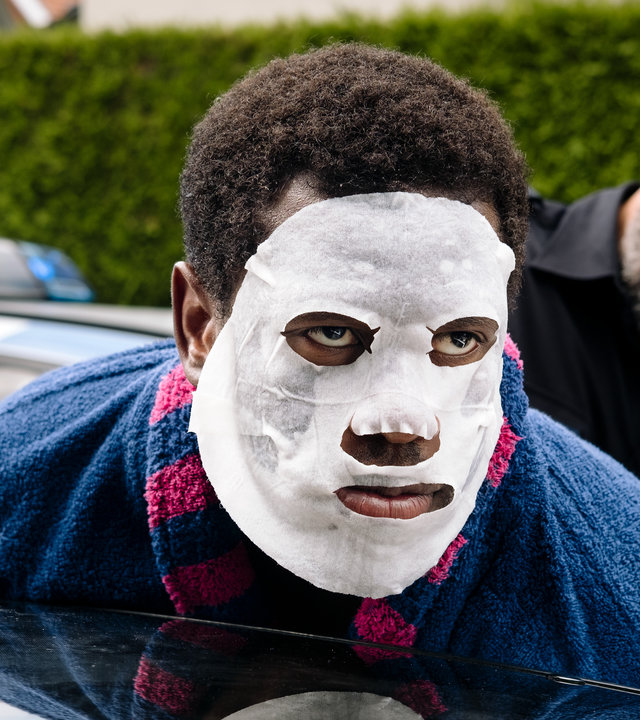 "I don't work here - Hecke": Dawit (Akeem van Flodrop) wird von Polizist Jakob (Robert Schupp) festgenommen. Während Jakob ihn in Polizeiuniform an der Autotür fixiert, lehnt Dawit seinen Oberkörper an ein dunkles Autodach und trägt eine weiße Tuchmaske und einen blauen Bademantel mit pinken Streifen.