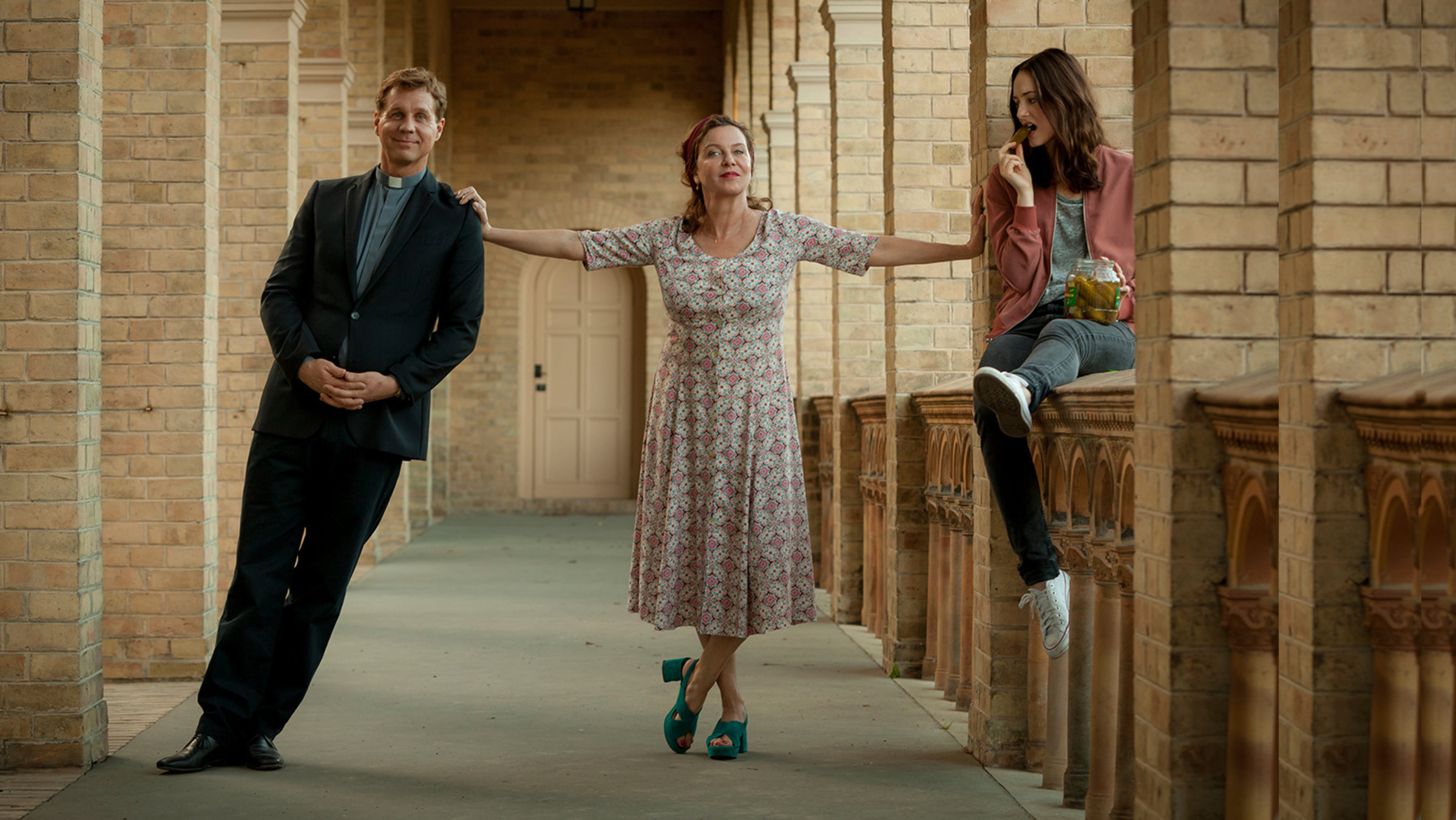 "Chaos-Queens: Ehebrecher und andere Unschuldslämmer": Pfarrer Benedikt (Thomas Heinze), Amelie (Margarita Broich) und Lou (Maria Ehrich) befinden sich in einem Kreuzgang. Benedikt lässt sich zur Seite fallen und wird von Amelie an der Schulter gehalten. Lou sitzt auf einer Brüstung, hält ein Gurkenglas in der Hand und isst eine Gurke. Sie schaut skeptisch zu ihrer Mutter.