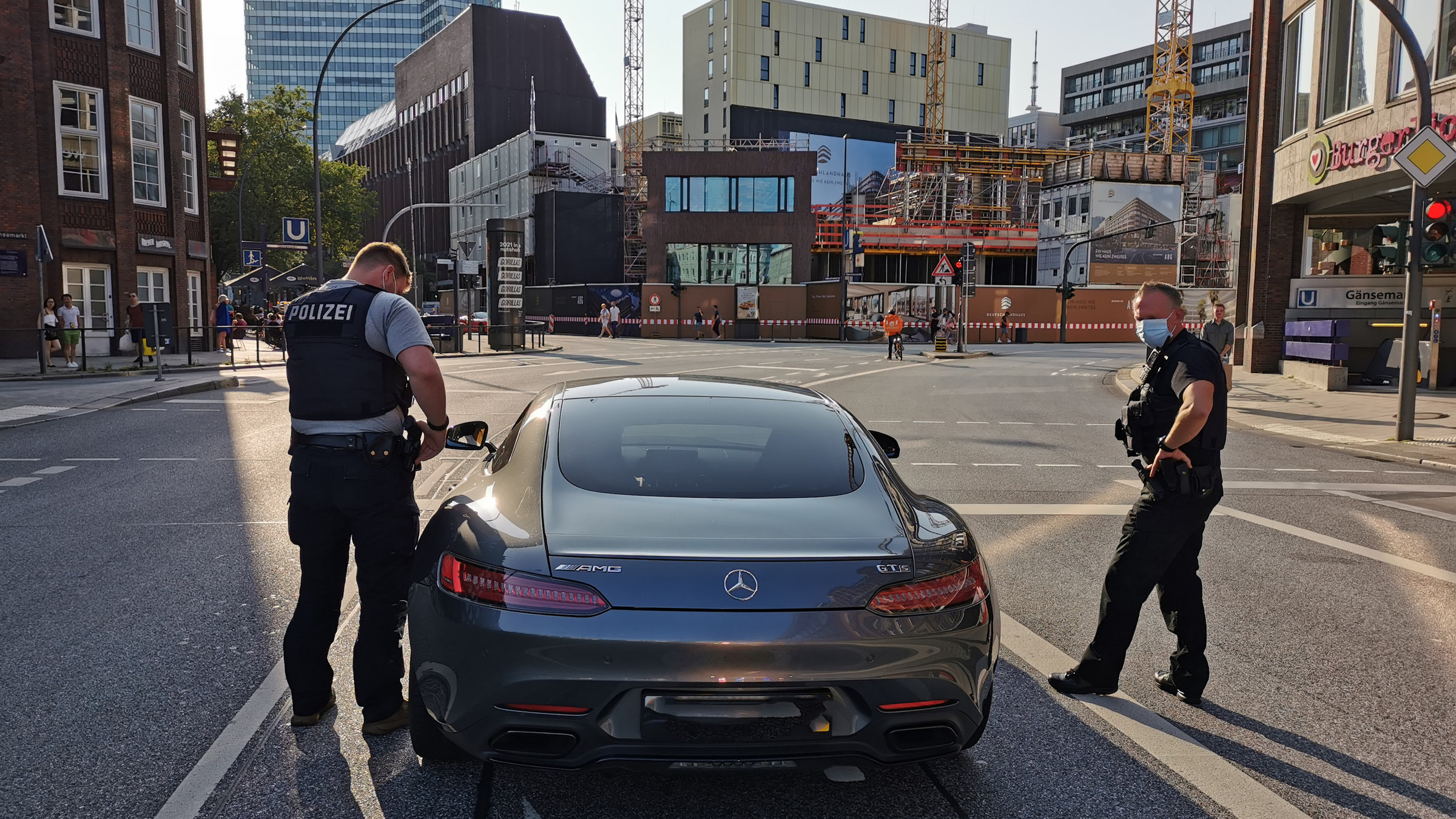 "ZDF.reportage: Viel PS und keine Rücksicht - Kampf gegen Autoposer": Eine Innenstadt-Kreuzung am späten Nachmittag, Blick von hinten auf einen Mercedes-Sportwagen. Zwei Beamte in zivil mit Polizeiweste stehen rechts und links vom Fahrzeug. Der linke Beamte prüft die Fahrzeugpapiere, der rechte die Reifen.