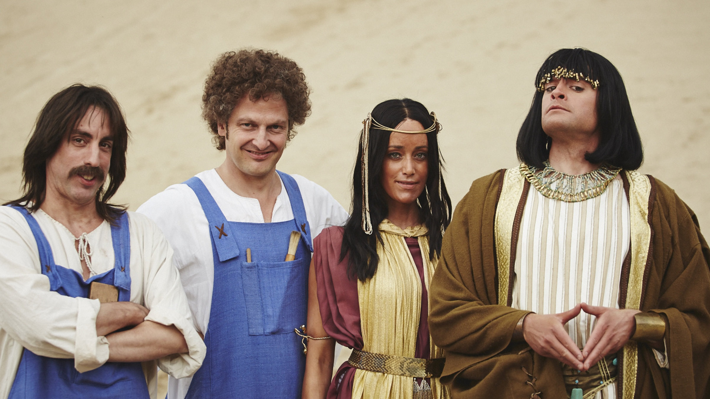 Alexander Schubert und Holger Stockhaus stehen als Bauunternehmer in blauen Togas verkleidet neben Judith Richter und Max Giermann, die in Gewänder des alten Ägypten gehüllt sind. Sie stehen in der Wüste und schauen direkt in die Kamera.