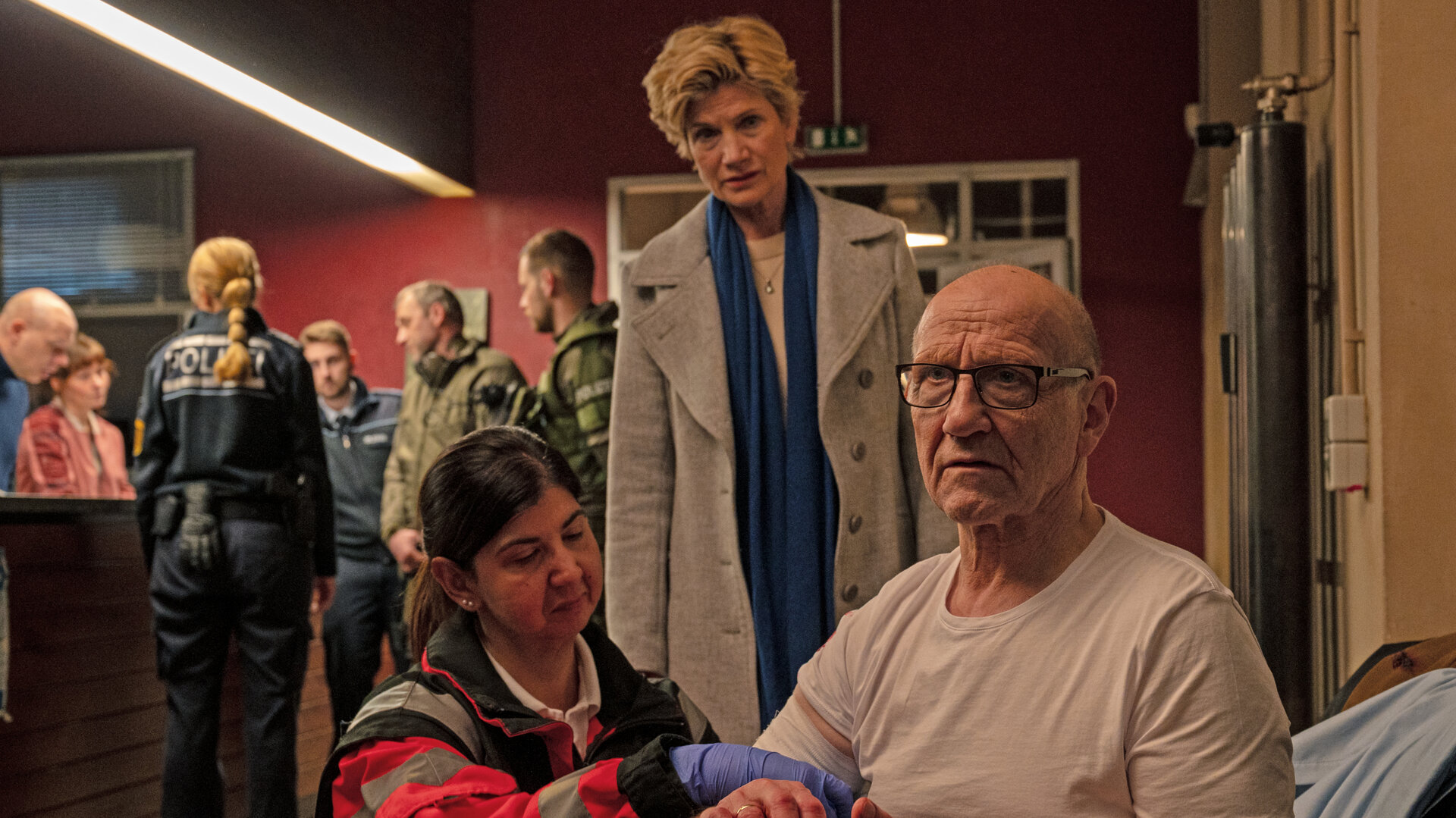 "SOKO Stuttgart - Kaiser im Visier": Kriminaldirektor Michael Kaiser (Karl Kranzkowski) bekommt neben der Wache im Präsidium von einer Notfallsanitäterin (Komparsin) einen Verband angelegt. Er schaut nachdenklich ins Leere. Hinter den beiden steht die Erste Kriminalhauptkommissarin Martina Seifert (Astrid M. Fünderich) und schaut Kaiser besorgt an. An der Wache im Hintergrund stehen Polizei- sowie SEK-Beamte und beraten sich.