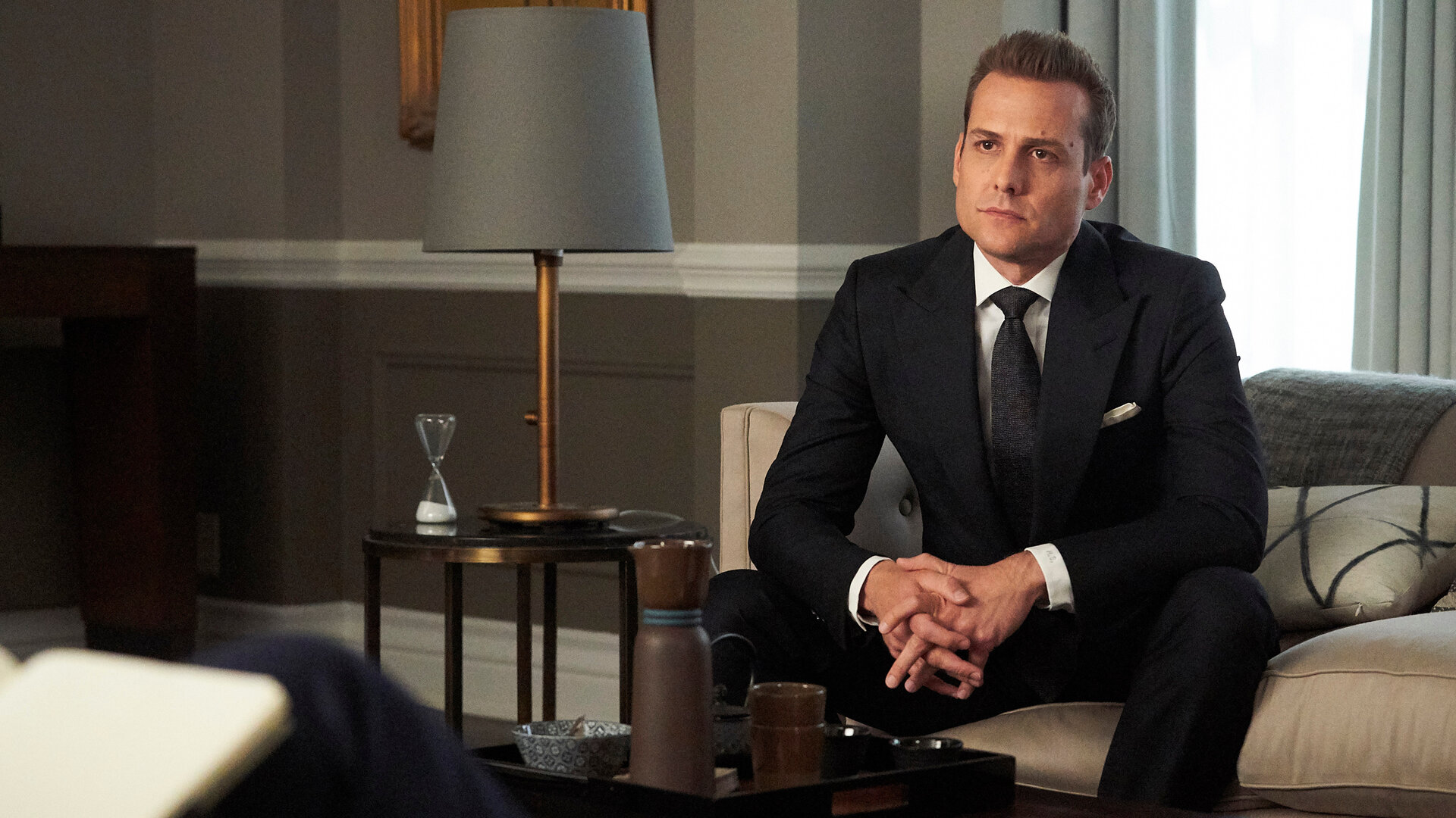 "Suits - Katzen, Ballett, Harvey Specter": Harvey (Gabriel Macht) - in einem dunklen Anzug - sitzt auf einem Sofa und schaut angespannt. Links neben ihm stehen auf einem Beistelltisch eine Lampe und eine Sanduhr.