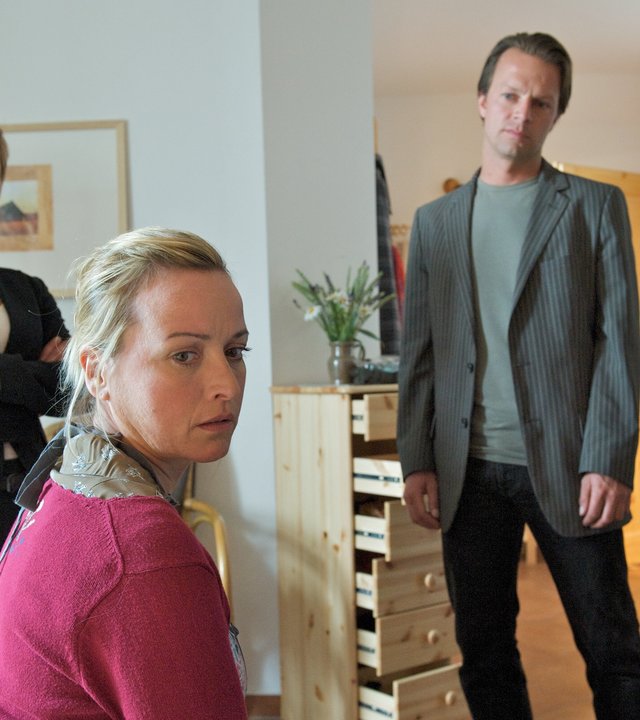 "SOKO Wismar - Frau Neumann": Irene Lüders (Katharina Abt) sitzt abgewandt von den Ermittlern im Zimmer von Frau Neumann, das von den Kommissaren Katrin Börensen (Claudia Schmutzler) und Sven Herzog (Michael Härle) durchsucht wurde. Die Schubladen einer Holzkommode stehen noch offen. Ihr Bruder Ralf Lüders (Thomas Arnold) steht im Flur und beobachtet die Szene.