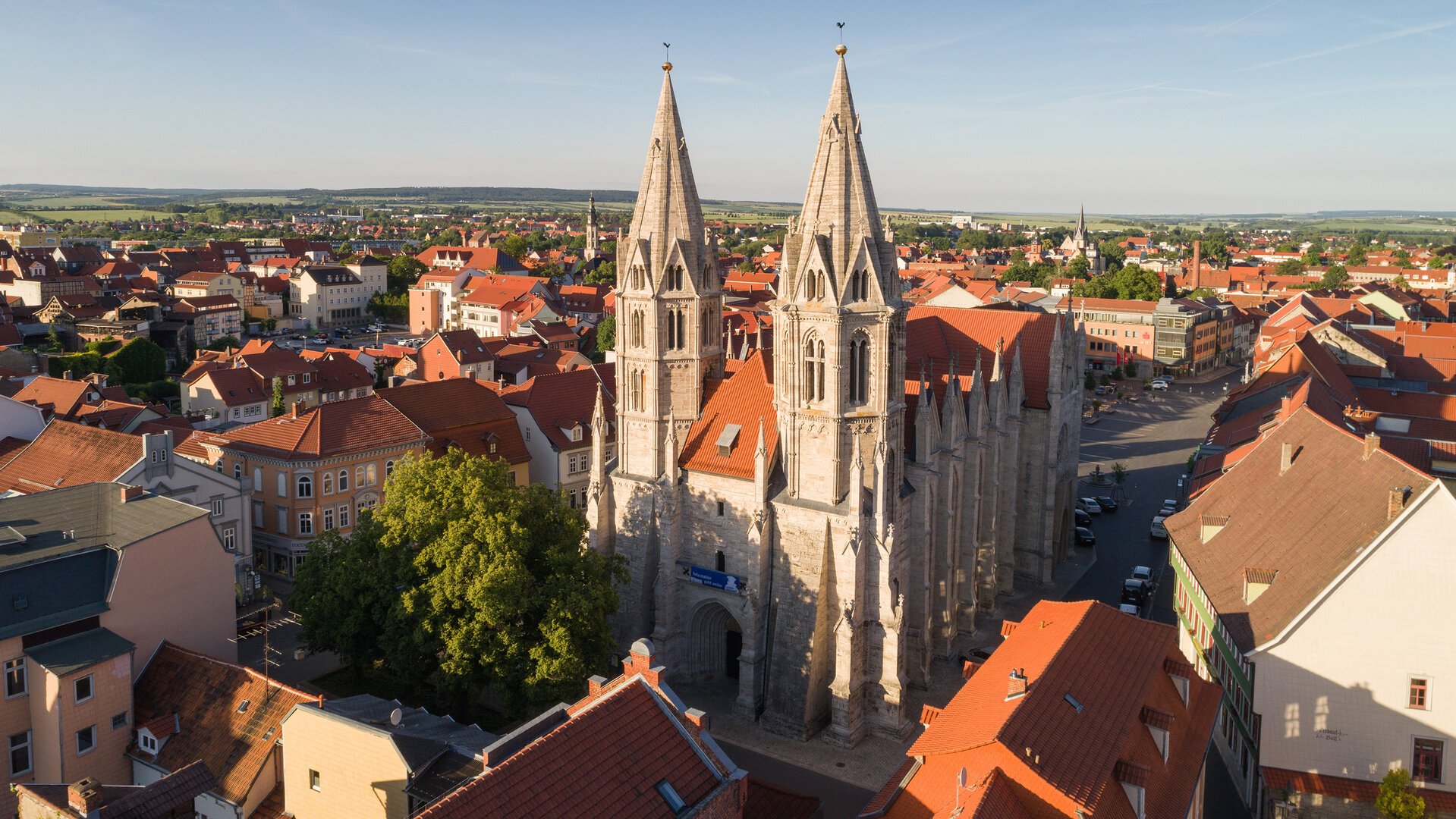 "Evangelischer Gottesdienst: 500 Jahre Bauernkrieg - vom Traum zur Katastrophe": Luftaufnahme: Die aus hellem Stein erbaute Divi-Blasii-Kirche liegt inmitten der Altstadt von Mühlhausen in Thüringen. Die beiden spitz zulaufenden Westtürme der Kirche ragen in den blauen Himmel. Im Hintergrund stehen Bäume vor sanften grünen Hügeln am Stadtrand von Mühlhausen.