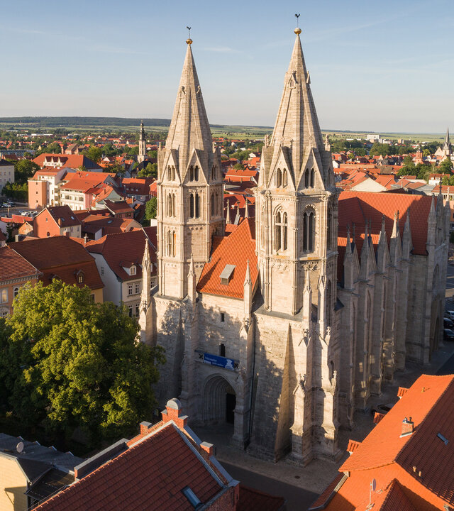 "Evangelischer Gottesdienst: 500 Jahre Bauernkrieg - vom Traum zur Katastrophe": Luftaufnahme: Die aus hellem Stein erbaute Divi-Blasii-Kirche liegt inmitten der Altstadt von Mühlhausen in Thüringen. Die beiden spitz zulaufenden Westtürme der Kirche ragen in den blauen Himmel. Im Hintergrund stehen Bäume vor sanften grünen Hügeln am Stadtrand von Mühlhausen.